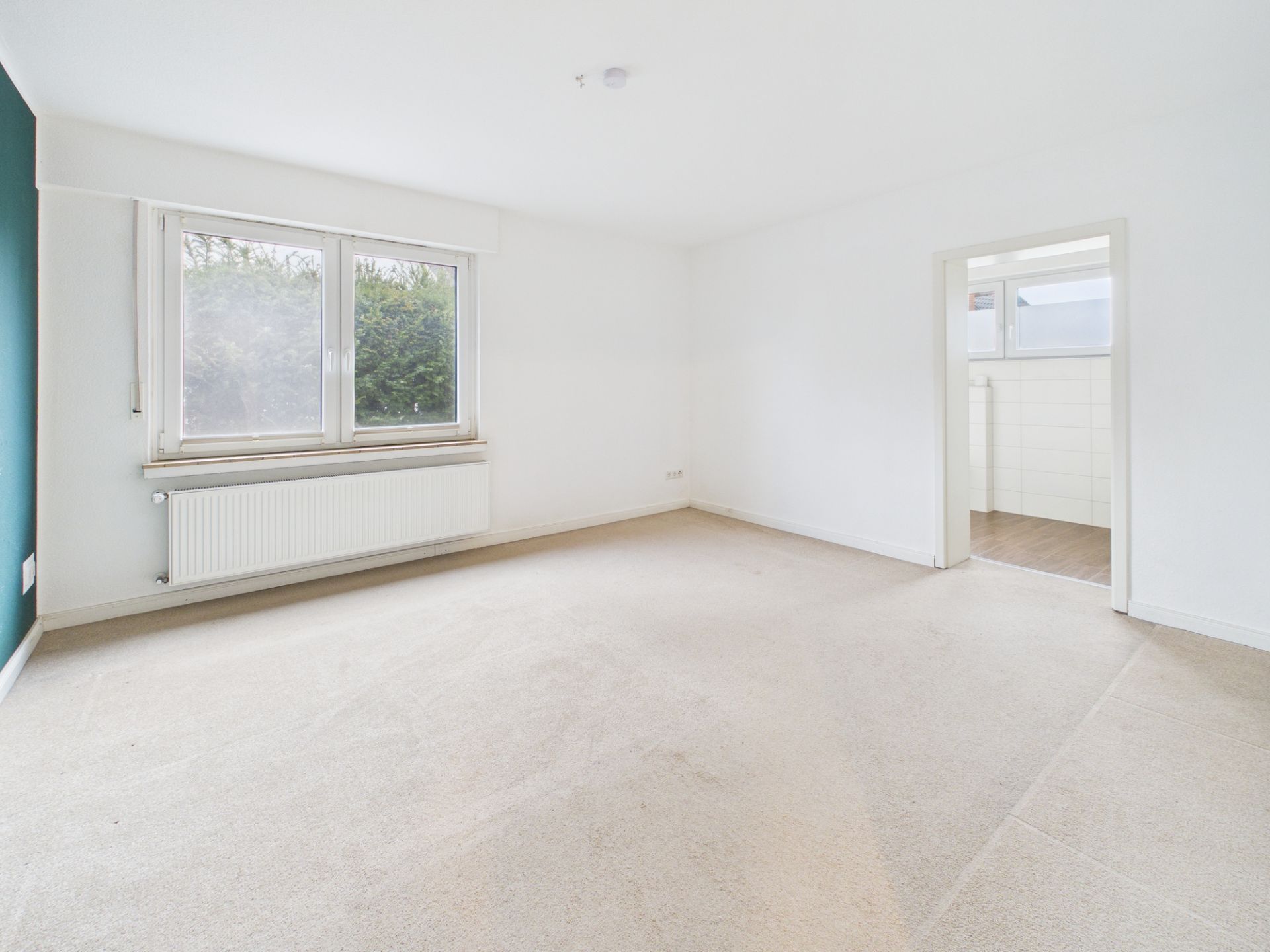 Helles, leeres Zimmer mit großem Fenster, Teppichboden und Zugang zu einem weiteren Raum.