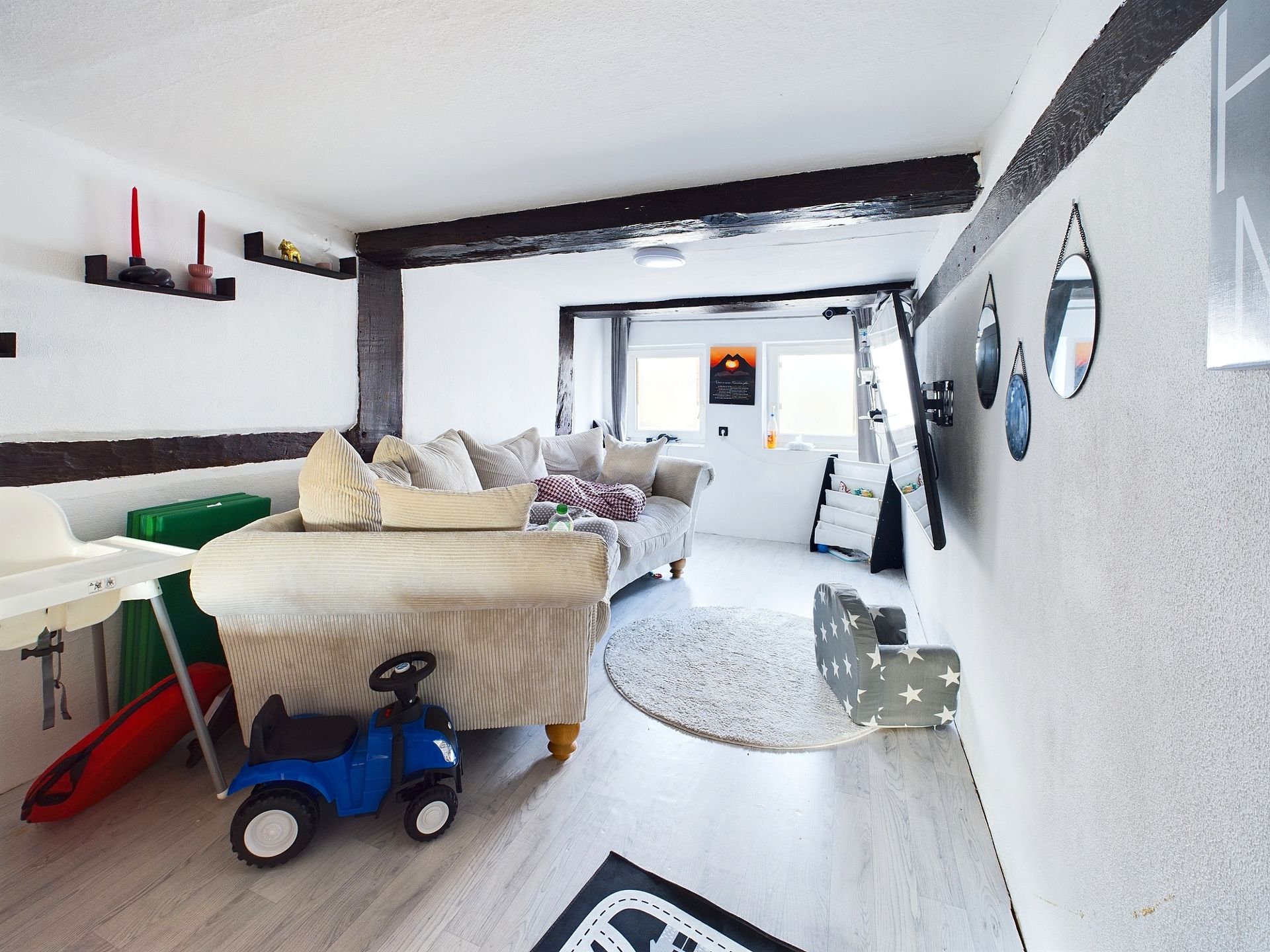Wohnzimmer mit Sofa, Spielzeugauto, Hochstuhl, Wandspiegeln und Spielmatte auf hellem Boden.