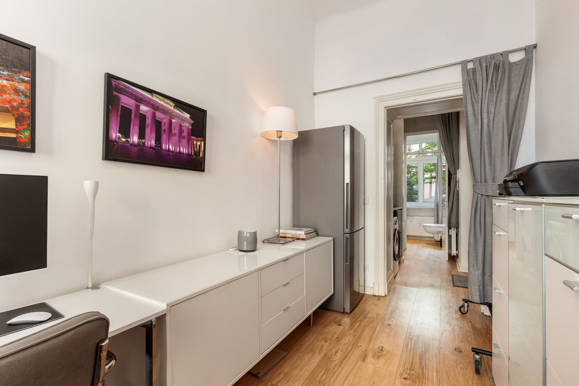 Heller Raum mit Schreibtisch, Stuhl, Kommode, Kühlschrank und Stehlampe, Blick auf einen Flur mit Fenster.