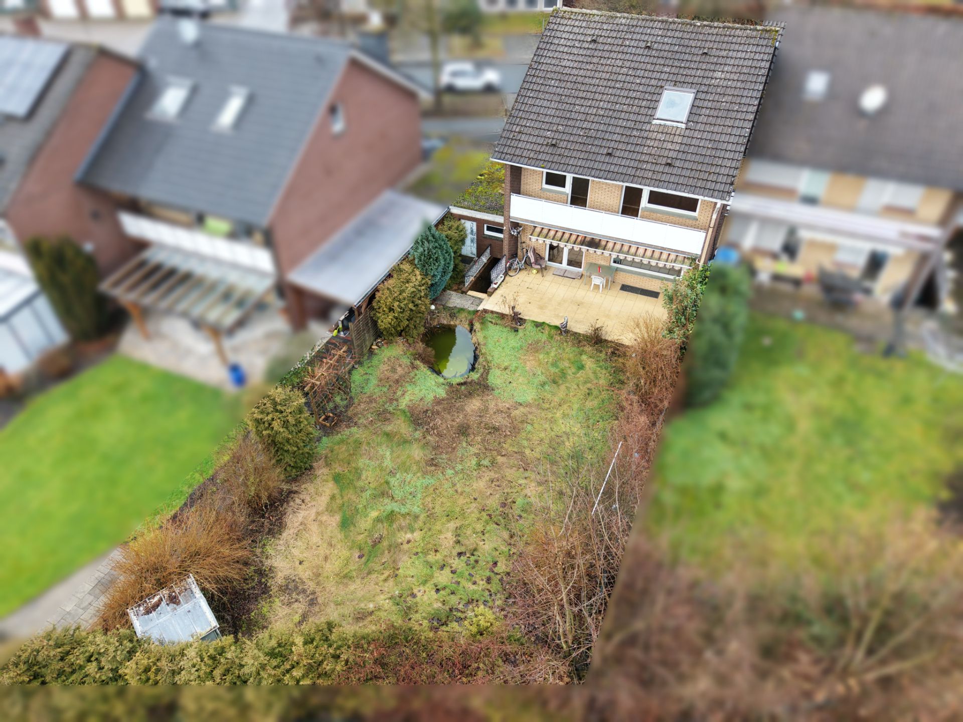 Luftaufnahme eines Gartens mit kleiner Terrasse, Teich und Wohnhaus im Hintergrund.