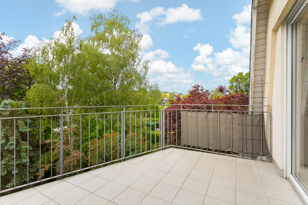 Heller Balkon mit Fliesenboden und Metallgeländer, umgeben von grünen Bäumen.