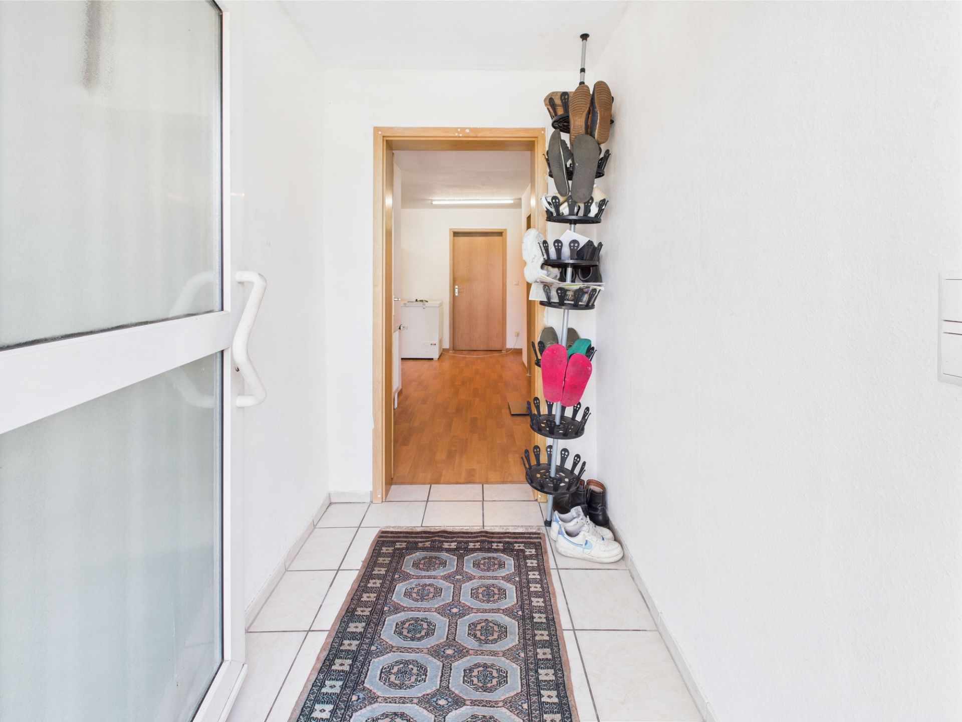 Heller Flur mit Teppich und Schuhregal an der Wand, Fliesenboden und offener Tür zum nächsten Raum.