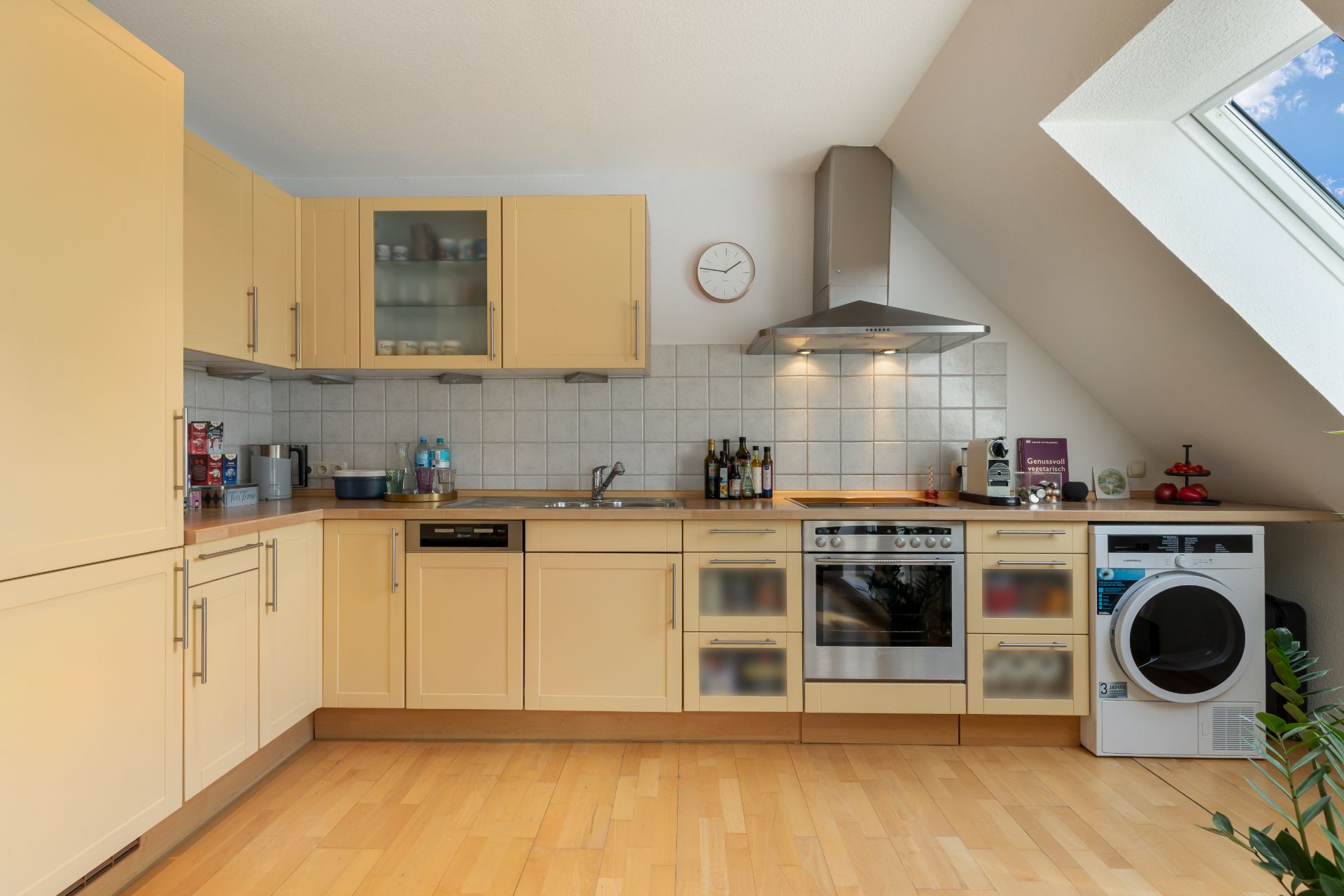 Moderne Einbauküche mit Herd, Spüle, Waschmaschine und großem Fenster unter einer Dachschräge.