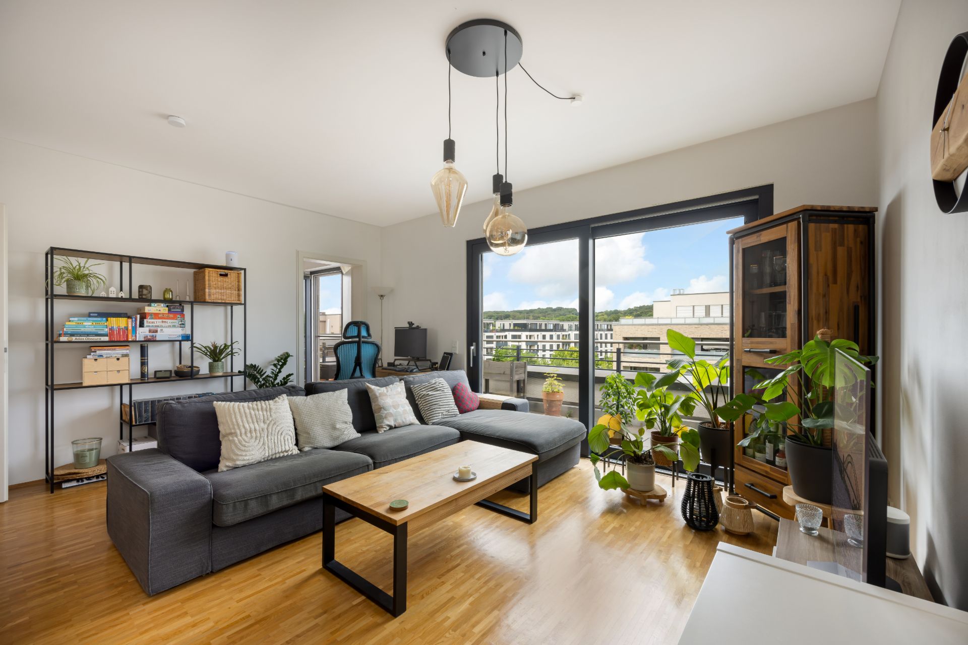 Helles Wohnzimmer mit grauem Sofa, Holztisch, Pflanzen und großem Fenster mit Blick ins Freie.