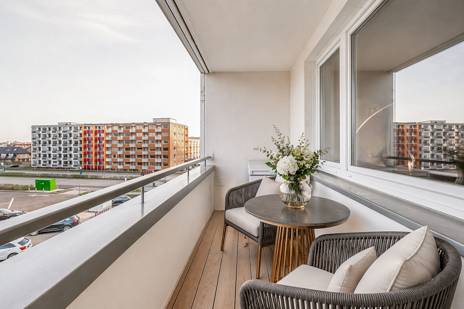 Balkon mit zwei Stühlen, rundem Tisch und Blumen in einer Vase, Ausblick auf Wohngebäude.