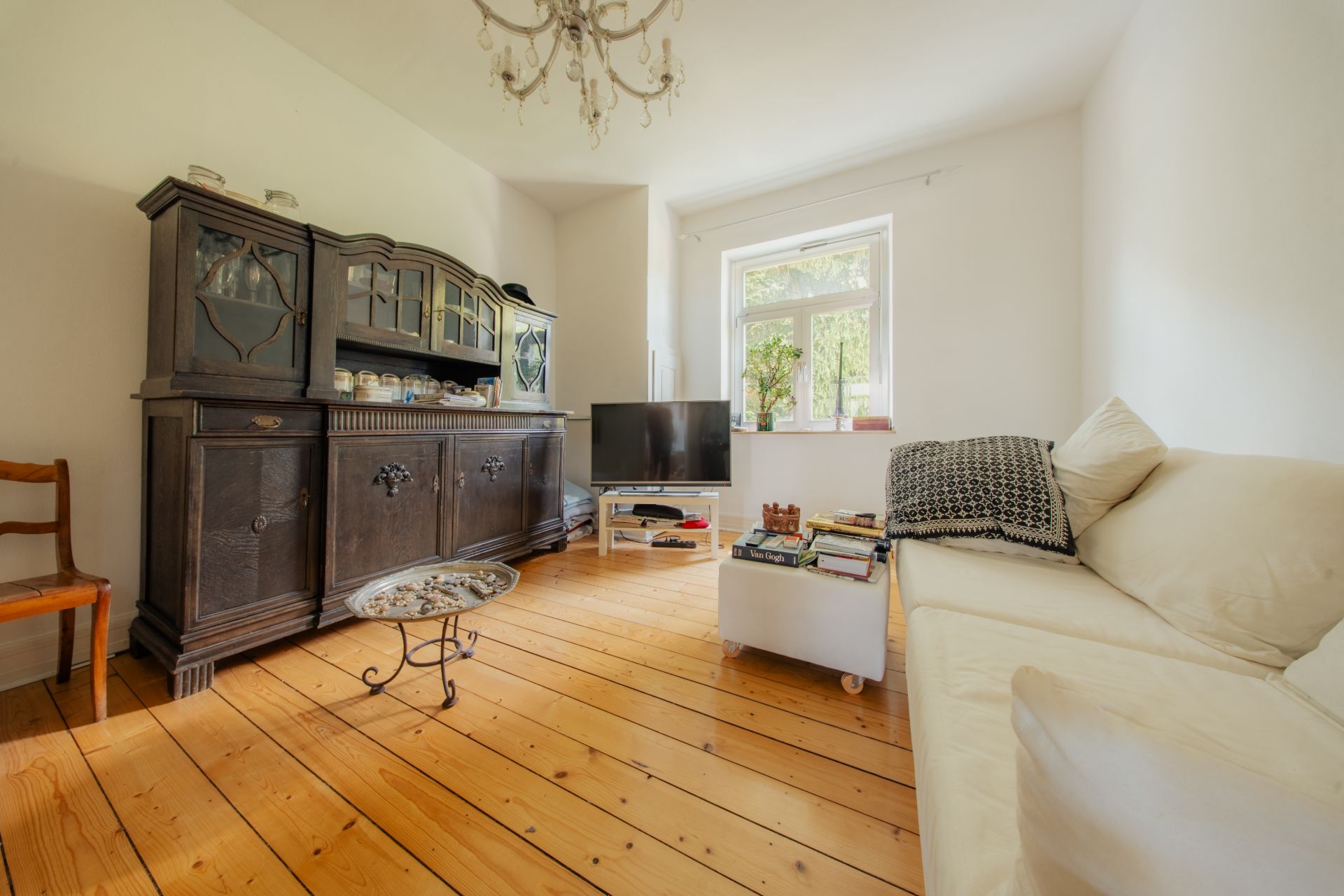Helles Wohnzimmer mit Holzfußboden, Sofa, dunklem Schrank, kleinem Tisch und Fenster.
