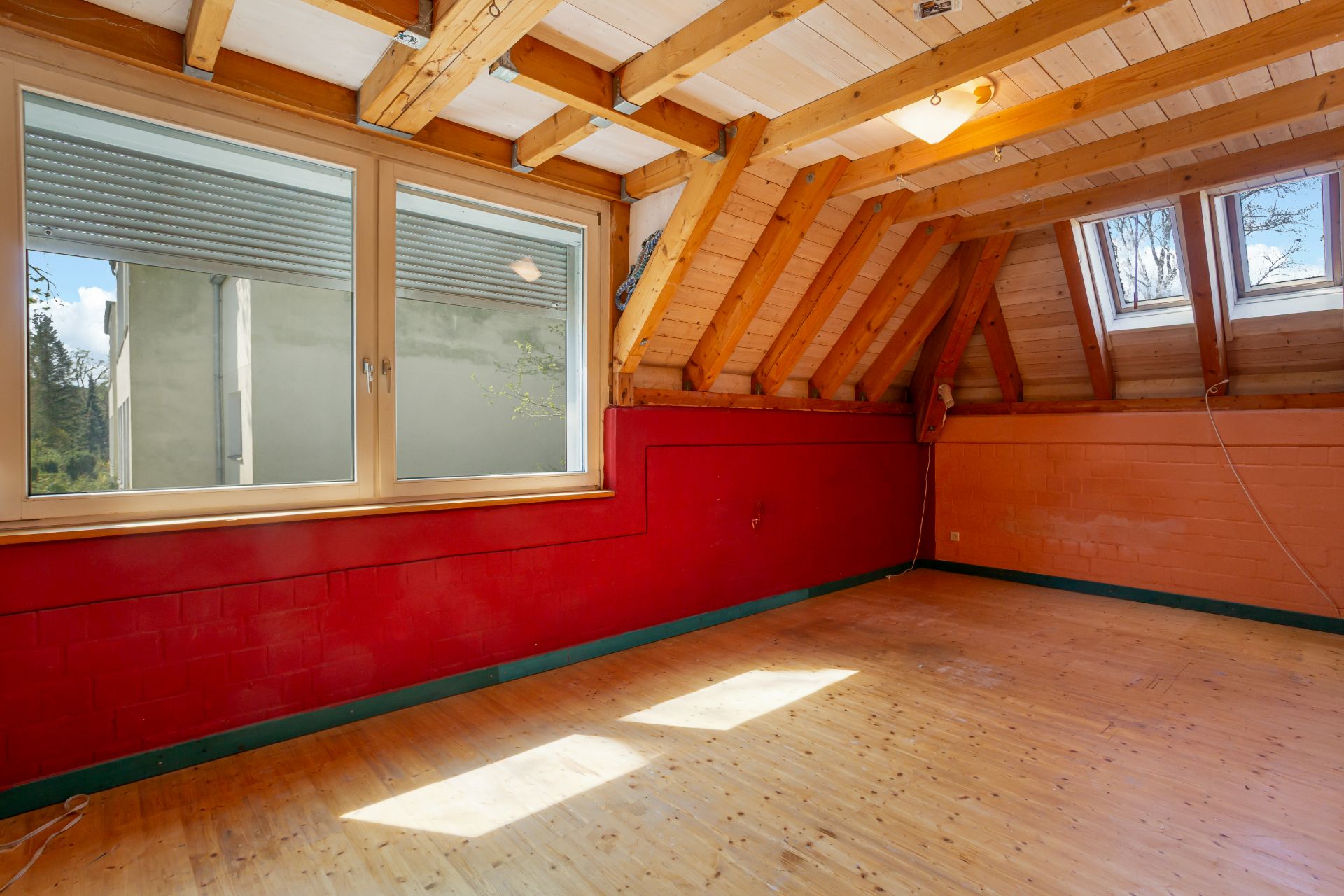 Dachzimmer mit Holzbalkendecke, großen Fenstern und roter Wand, Holzboden ohne Möbel.