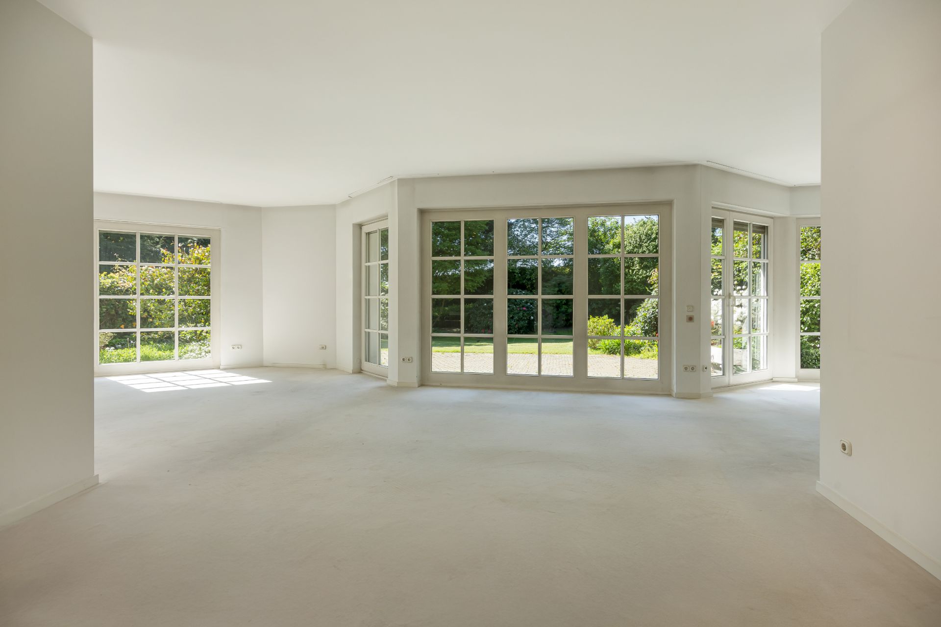 Helles, leeres Zimmer mit großen Fenstern und Blick auf einen grünen Garten.