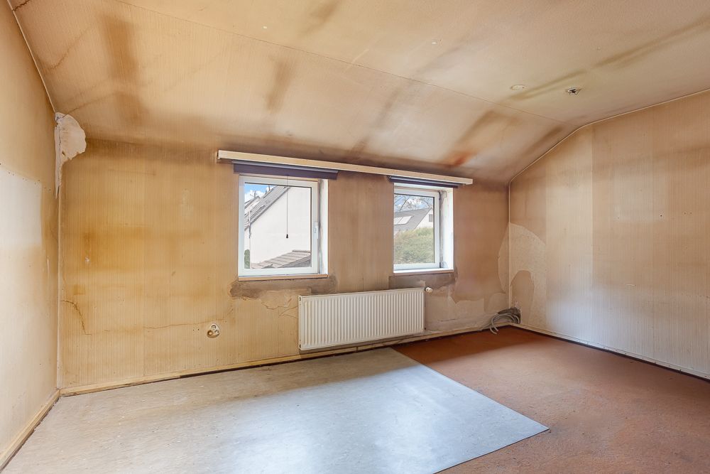 Leerer Raum mit zwei Fenstern, altem Bodenbelag und abgenutzten Wänden unter einer Dachschräge.