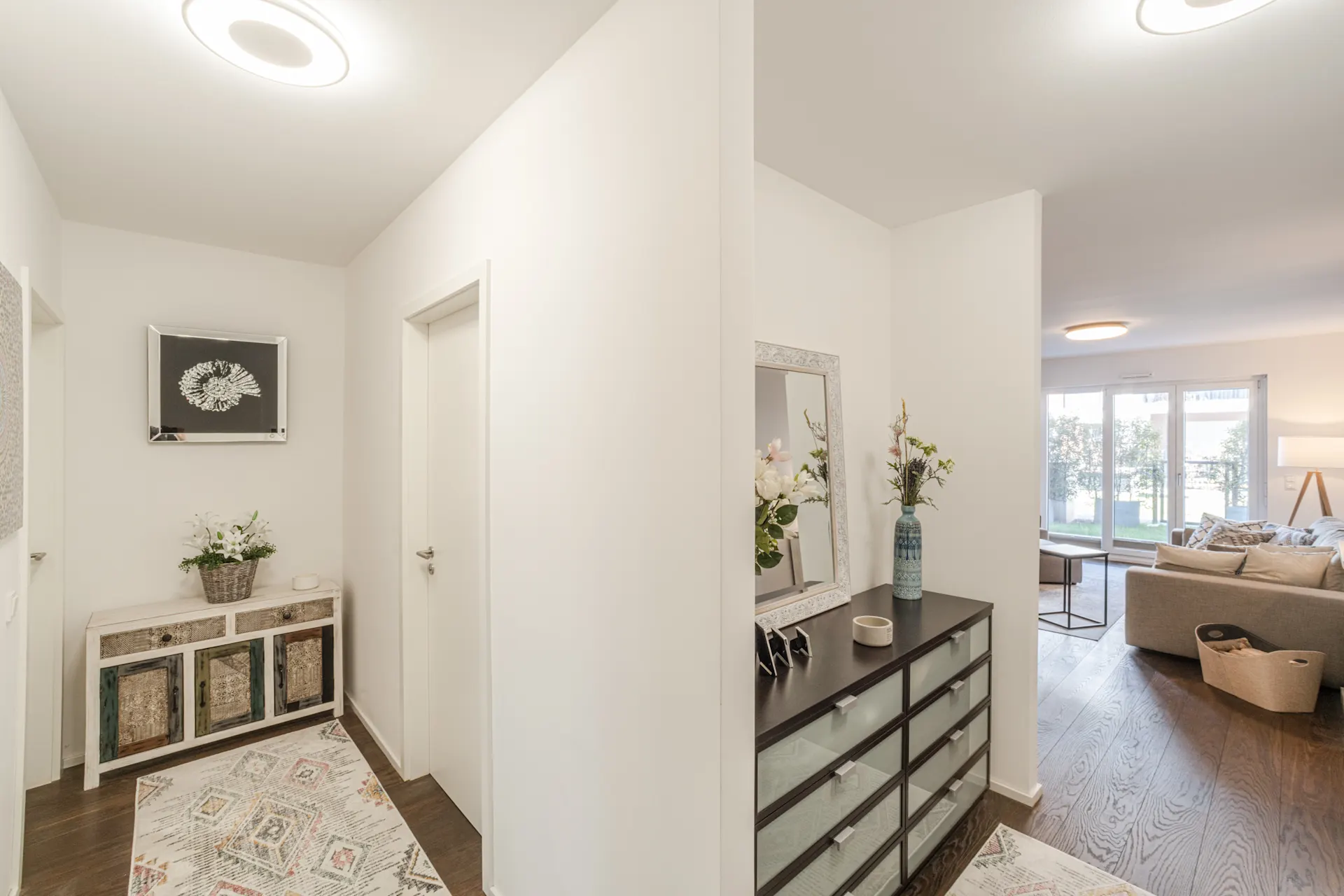 Heller Flur mit Kommode, Teppich, Blumen und Blick ins Wohnzimmer mit Sofa.