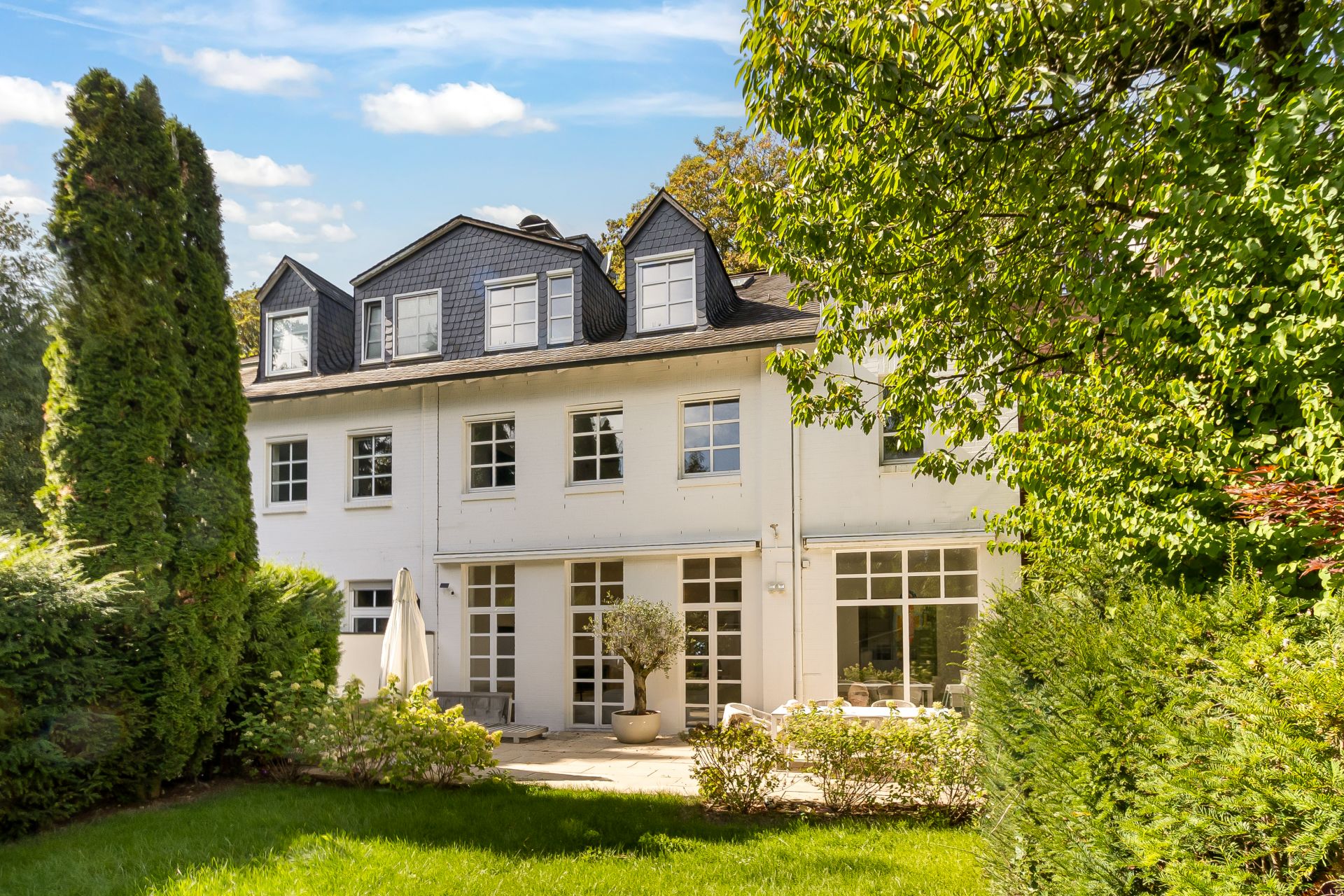 Helles Wohnhaus mit Garten, großen Fenstern und Terrasse, umgeben von Bäumen und Sträuchern.