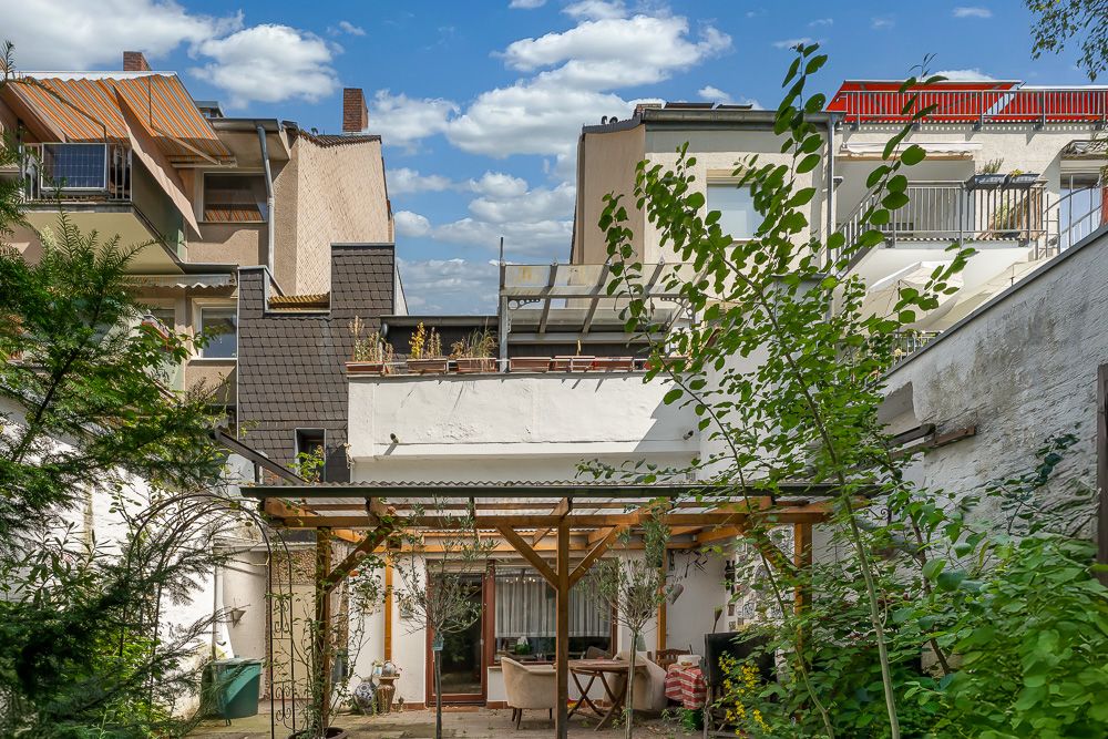 Hinterhofgarten mit Pergola, Pflanzen und angrenzendem Gebäude im Wohnbereich bei Tageslicht.