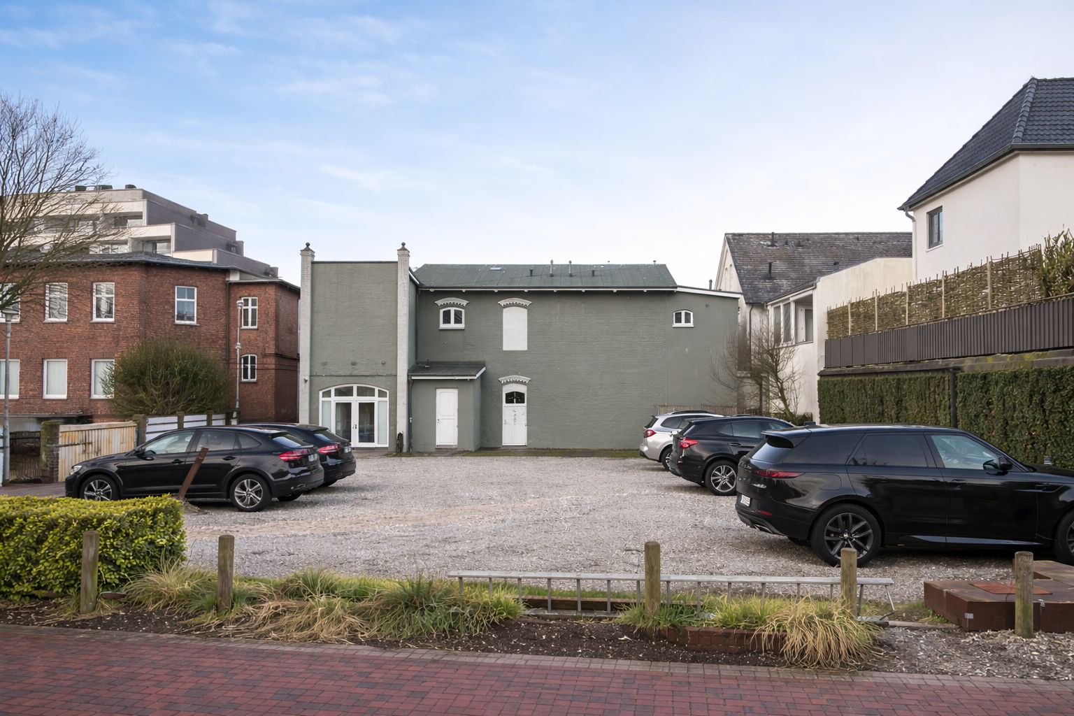 Mehrere Autos parken auf einem Kiesparkplatz vor einem grauen Wohnhaus.