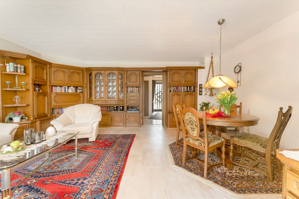Wohnzimmer mit Holzmöbeln, Teppichen, rundem Esstisch und dekorativen Blumenvasen.