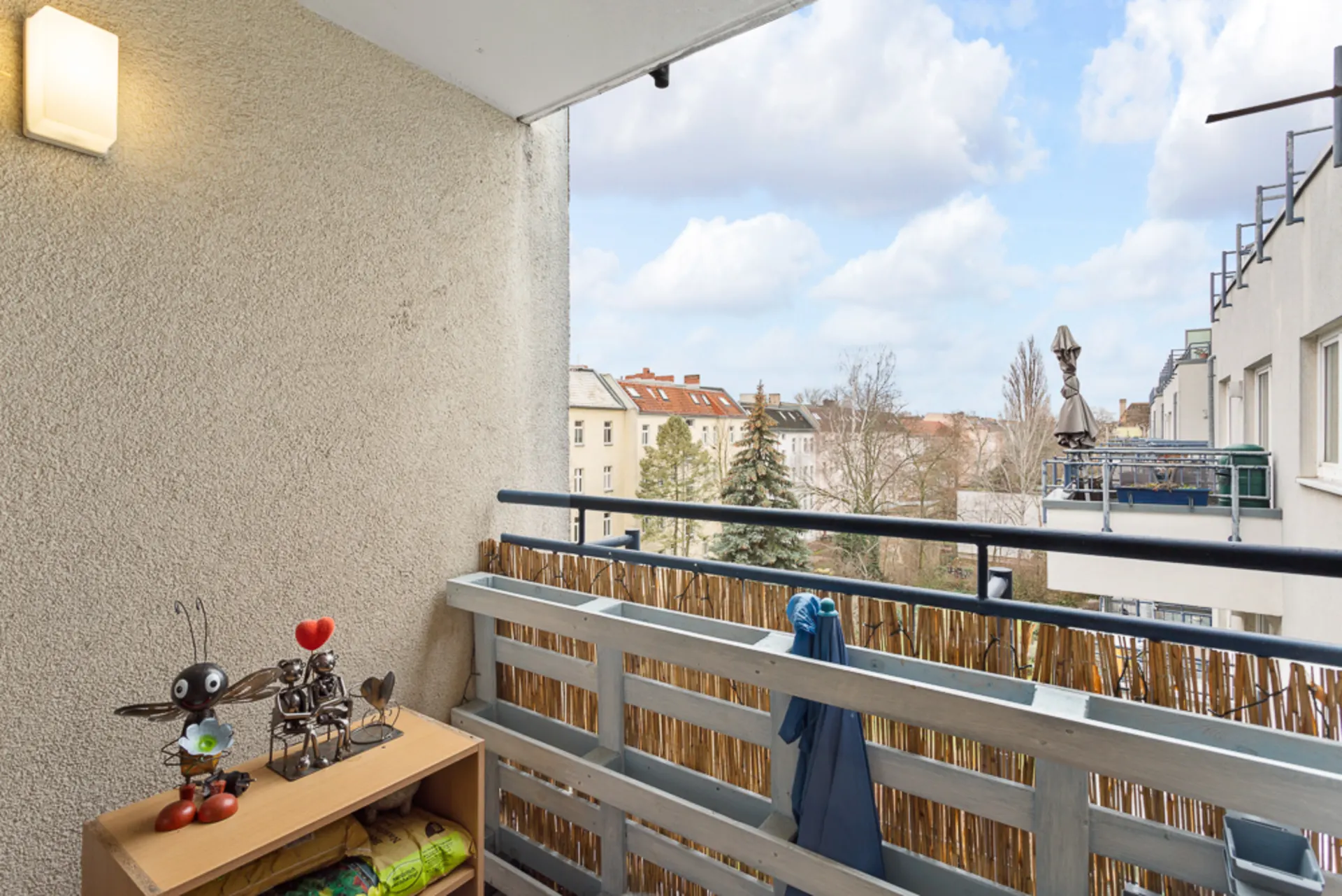Kleiner Balkon mit Holzgeländer, Regal und Blick auf Bäume und Nachbarhäuser.