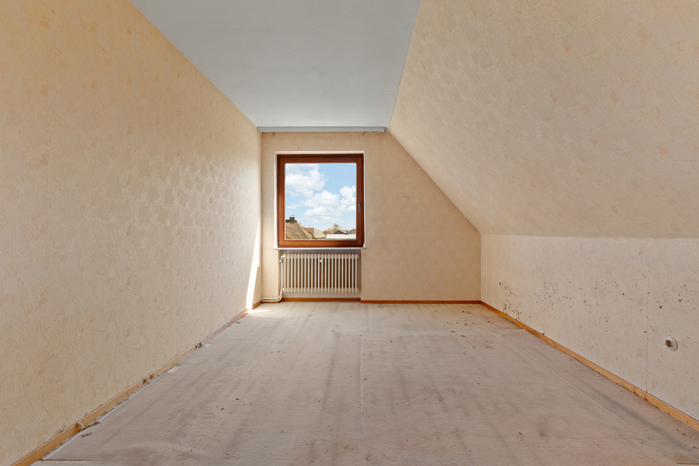 Leerer Raum mit Dachschräge, hellem Teppich und Fenster in der Mitte der Stirnwand.