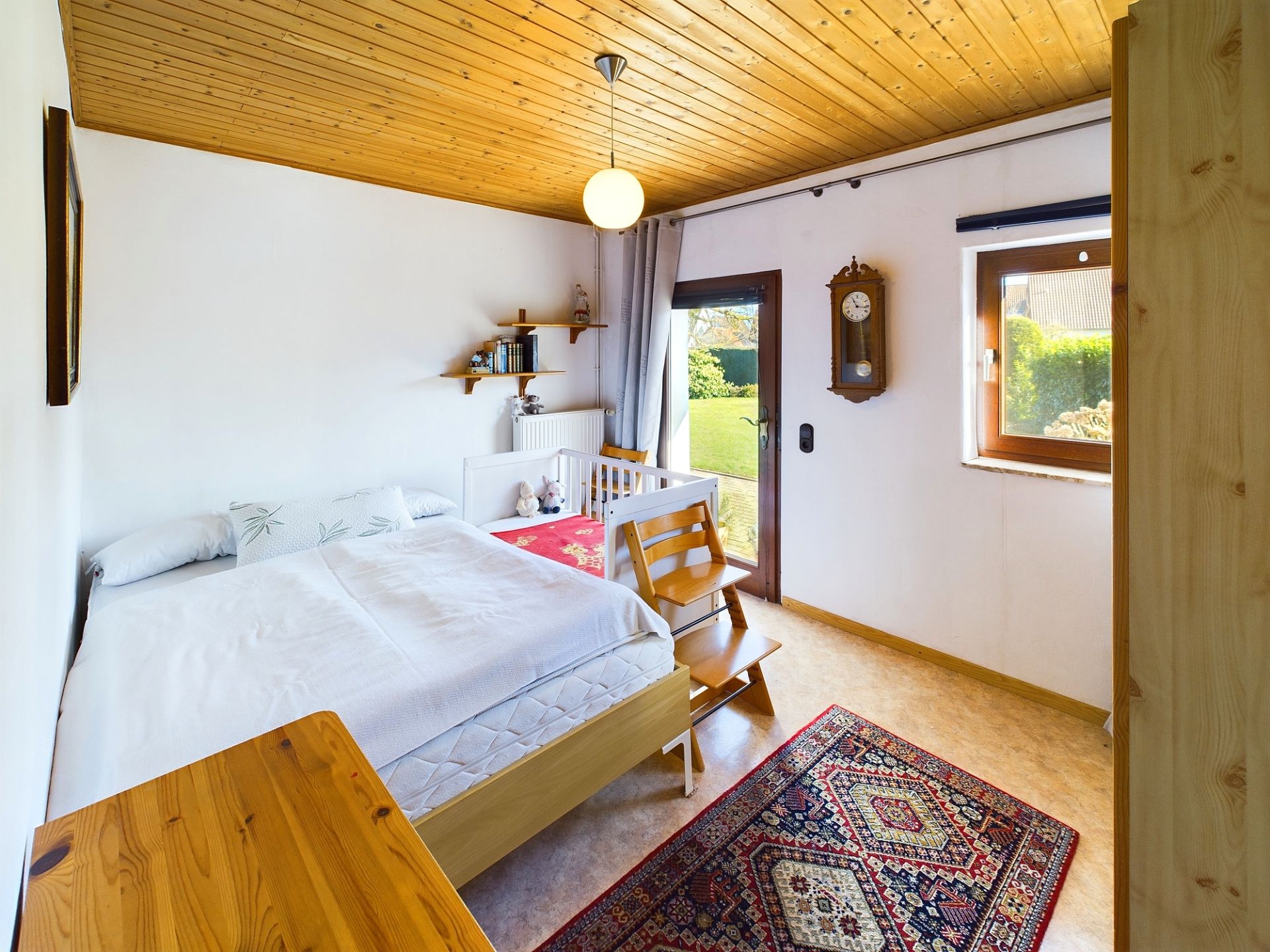 Helles Schlafzimmer mit Doppelbett, Kinderbett, Teppich, Holzmöbeln und Zugang zum Garten.