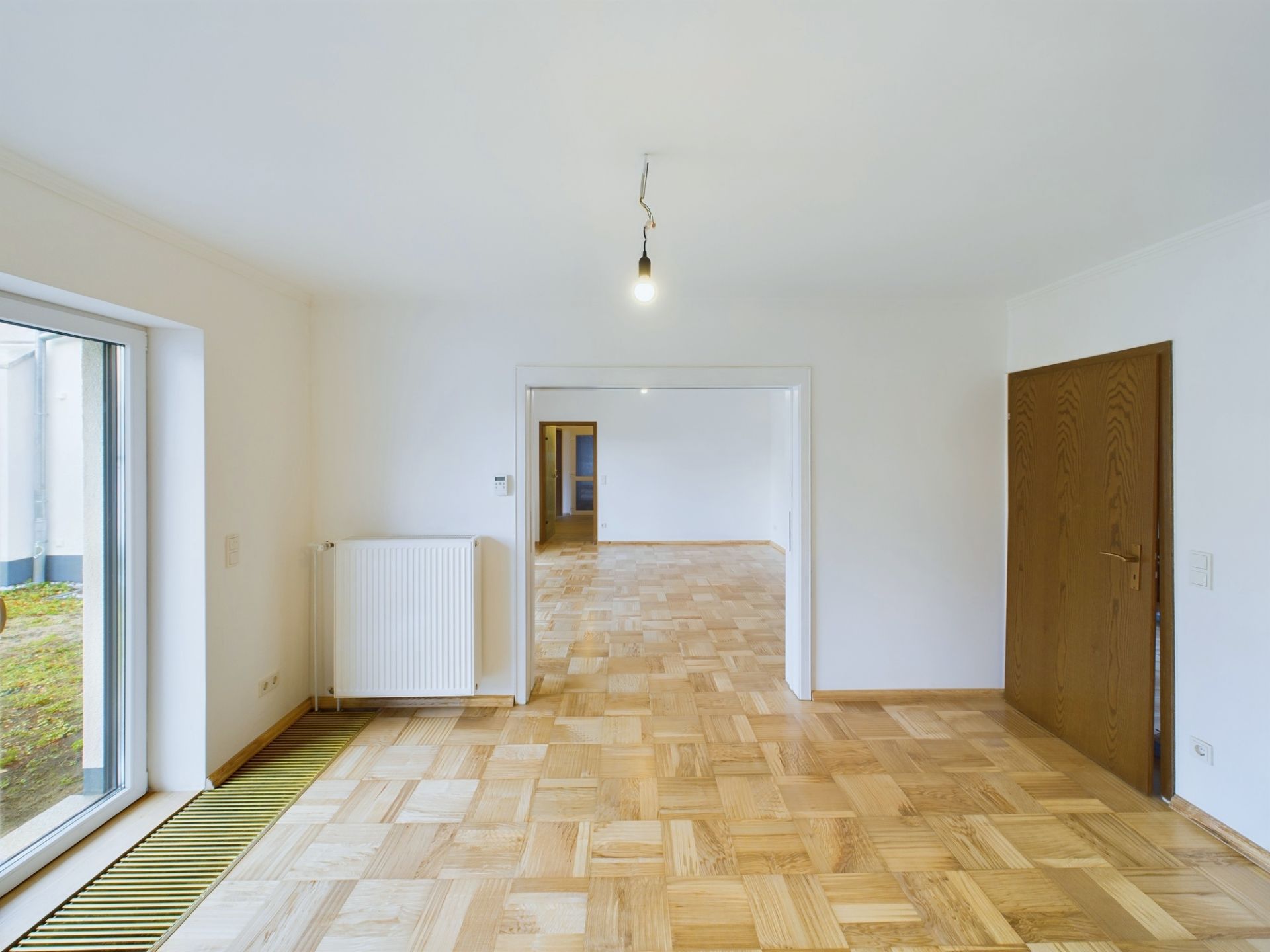 Helles Zimmer mit Parkettboden, großer Türöffnung, Fenster, Heizkörper und brauner Tür.