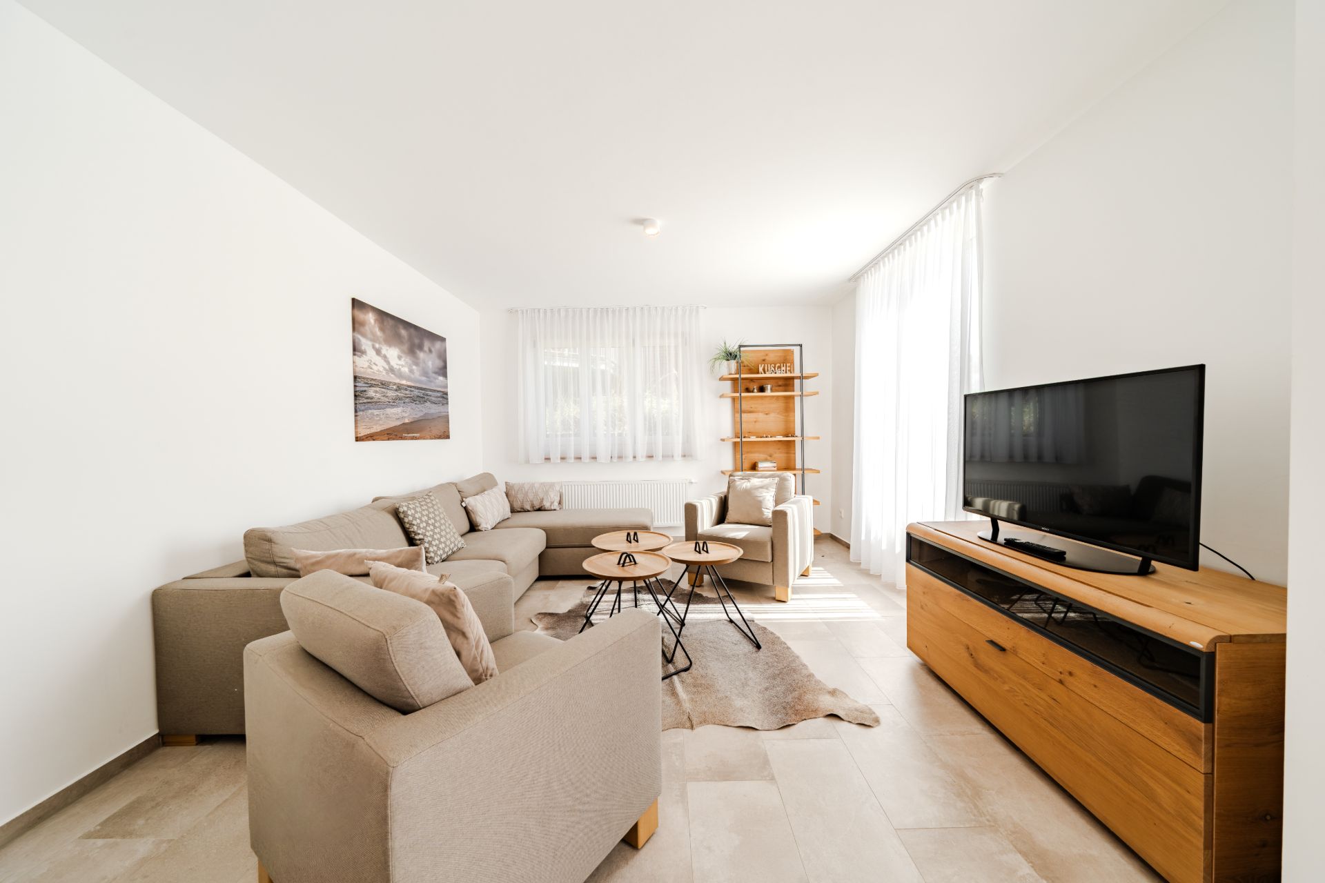 Helles Wohnzimmer mit beigen Sofas, TV, Holzregal und Couchtischen, große Fenster mit weißen Vorhängen.
