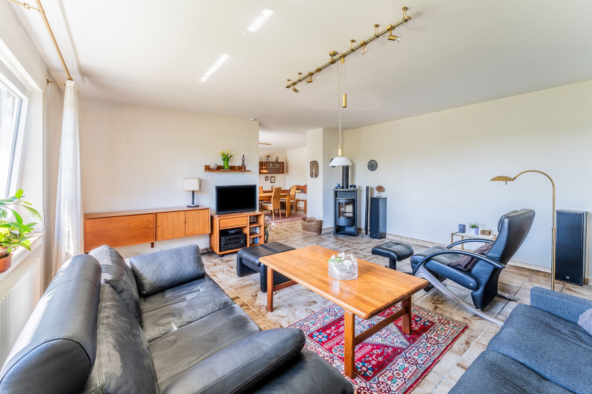 Helles Wohnzimmer mit Ledersofas, Holztisch, TV, Kamin und Esstisch im Hintergrund.