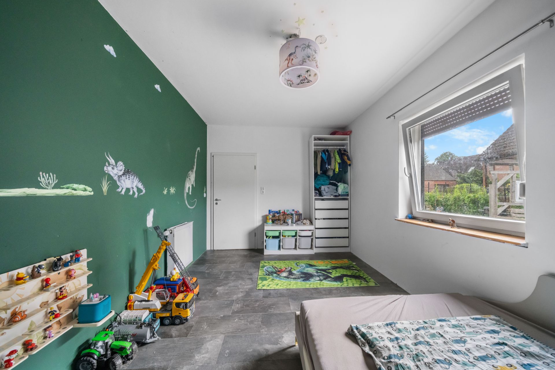 Kinderzimmer mit grün gestrichener Wand, Spielzeug, Bett, Fenster und Wandregalen.