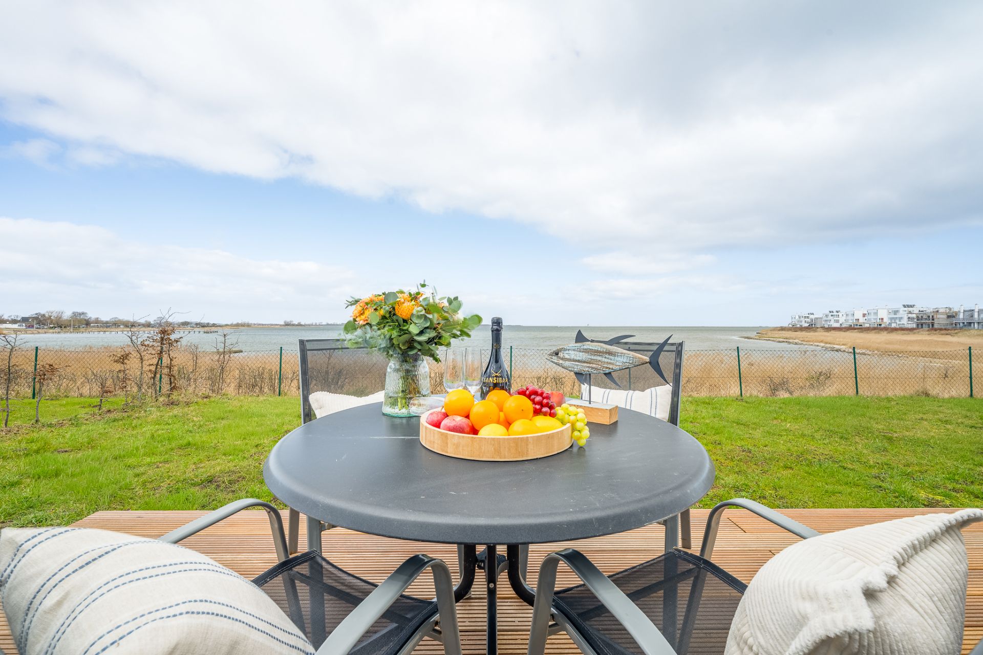 Runder Terrassentisch mit Obst, Blumen und Wein, Blick auf Garten und Wasser im Hintergrund.