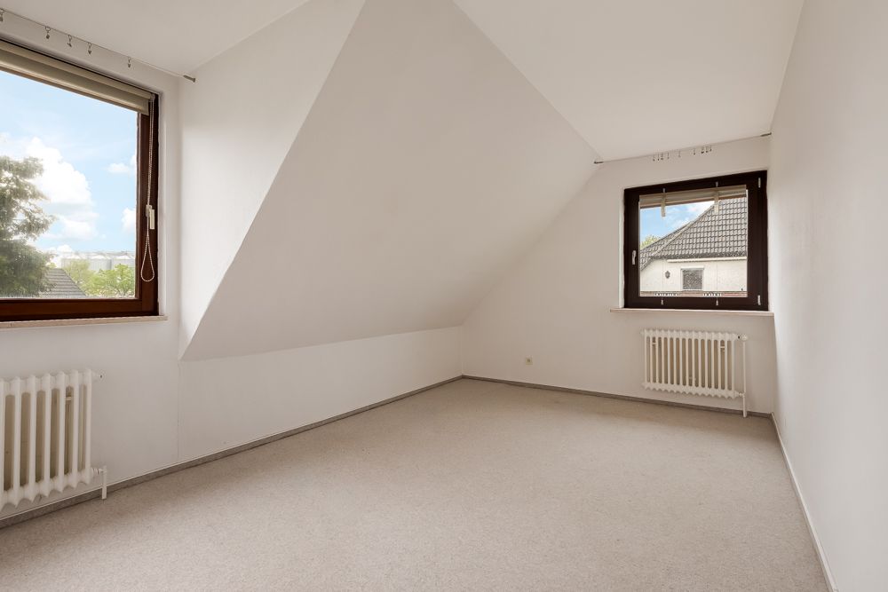 Helles, leeres Zimmer mit zwei Fenstern, weißen Wänden und Dachschräge.