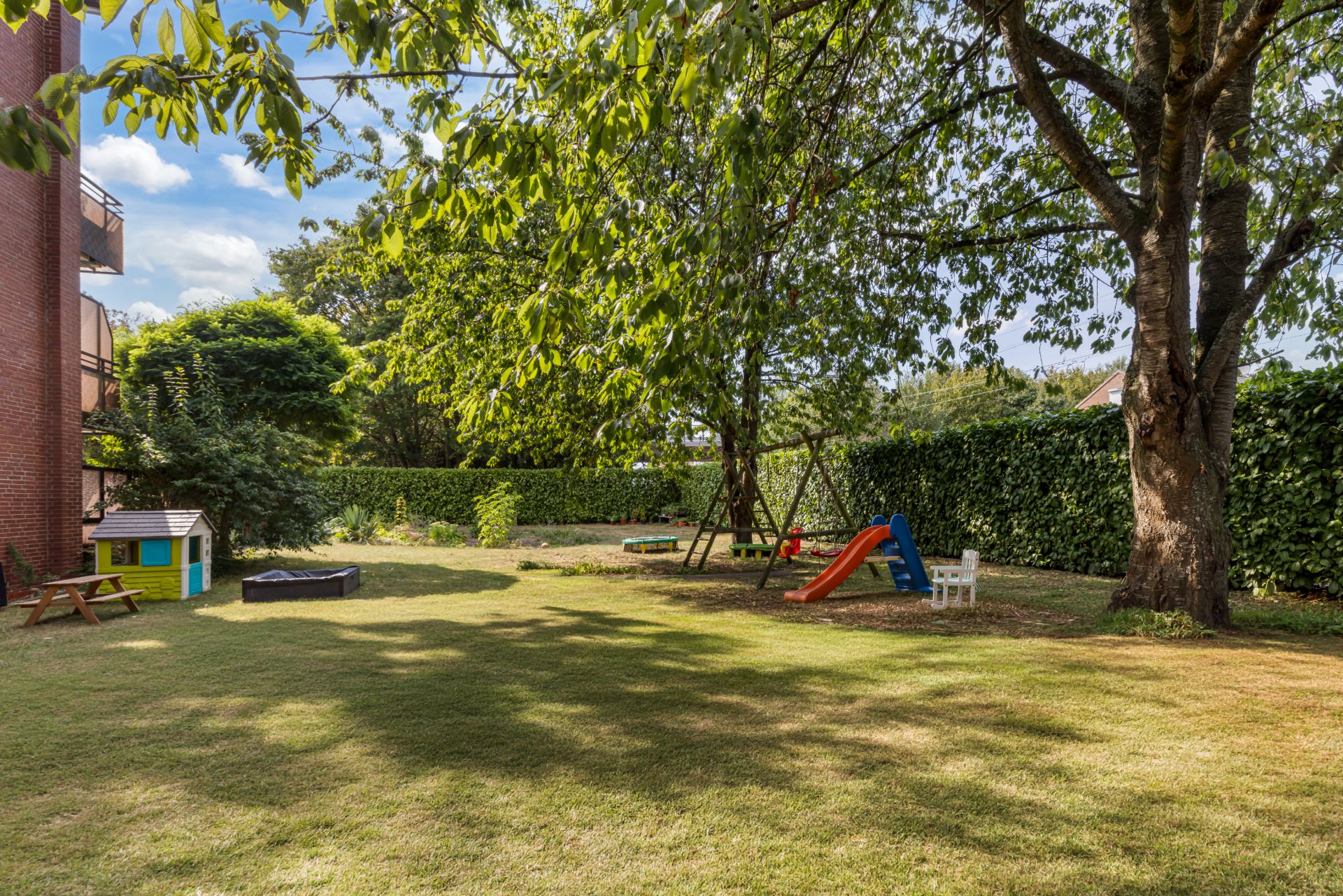 Garten mit Spielhaus, Rutsche, Schaukel und großen Bäumen auf einer Rasenfläche.