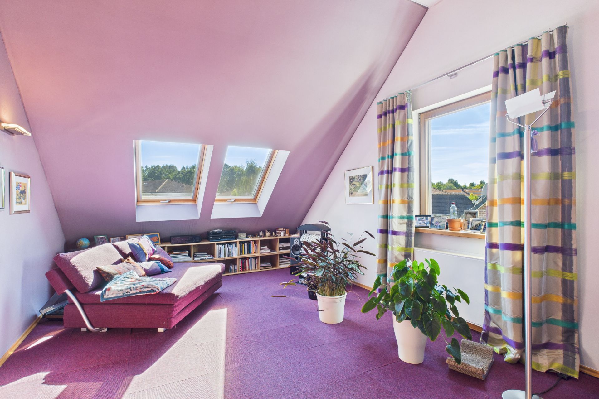 Helles Zimmer mit schräger Decke, Sofa, Bücherregal, Pflanzen und großen Fenstern.