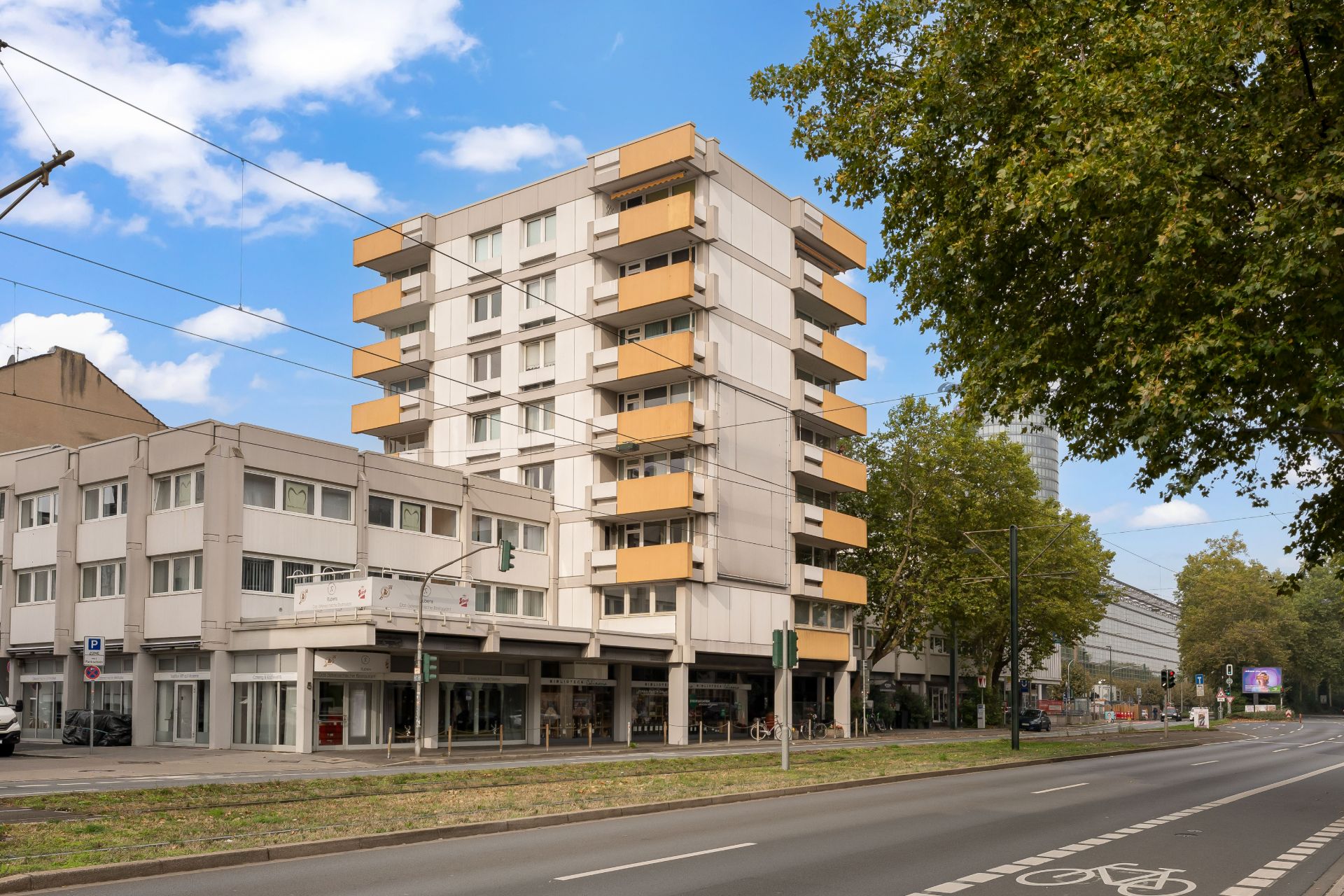 Mehrstöckiges Wohnhaus mit gelben Balkonen an einer Straße, Bäume und Geschäfte im Erdgeschoss.