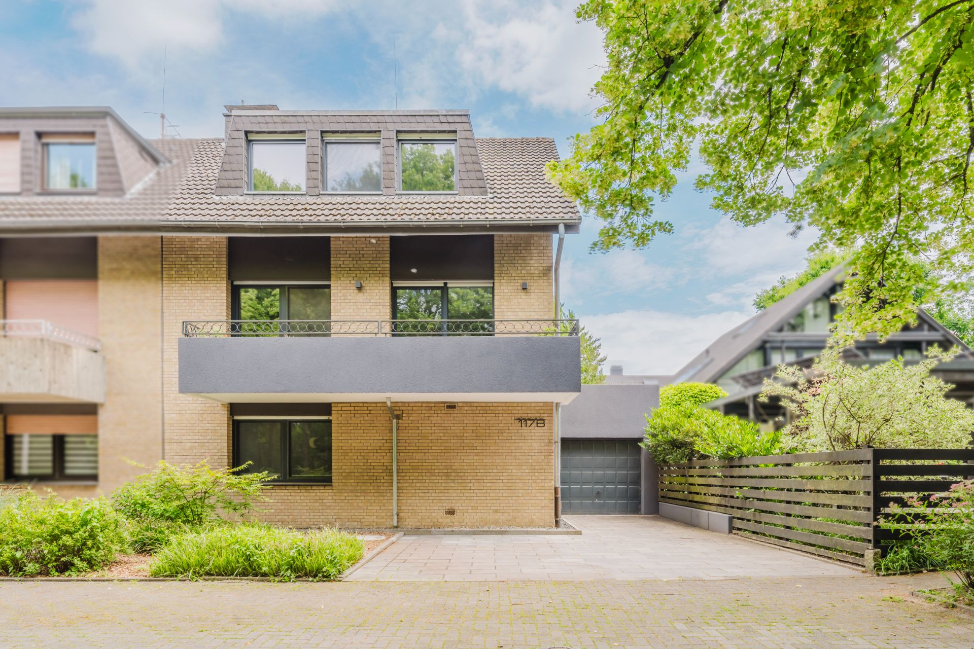 Moderne, dreistöckige Wohnhaus-Fassade mit Garage, Balkon und Auffahrt, umgeben von Bäumen und Zaun.