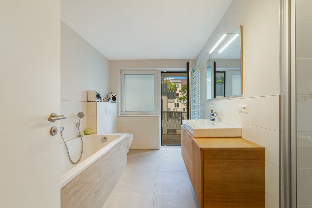 Helles Badezimmer mit Badewanne, Waschbecken, Spiegel und Fenster zum Balkon.