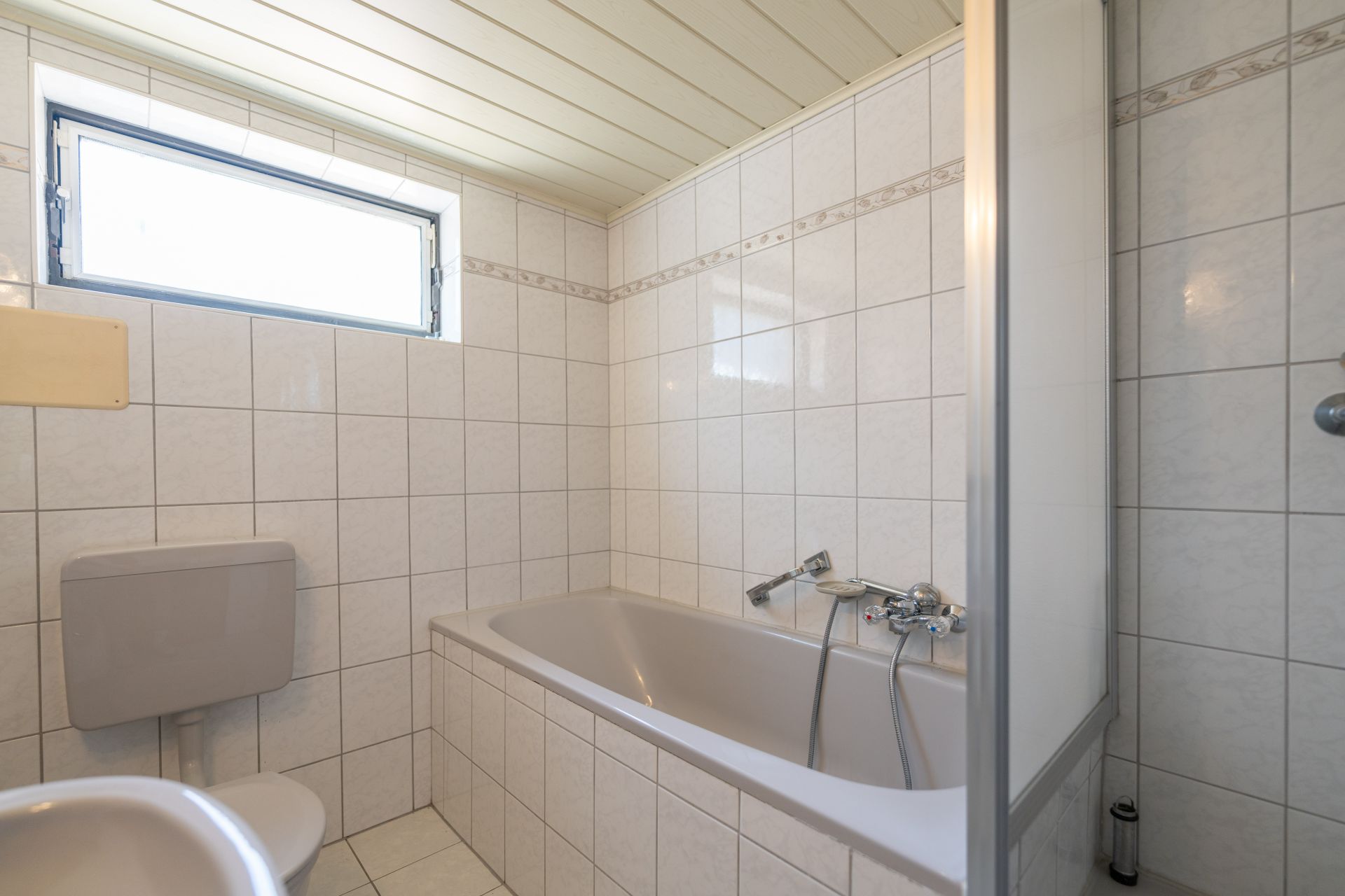 Badezimmer mit Badewanne, Toilette und kleinem Fenster, alles mit weißen Fliesen versehen.