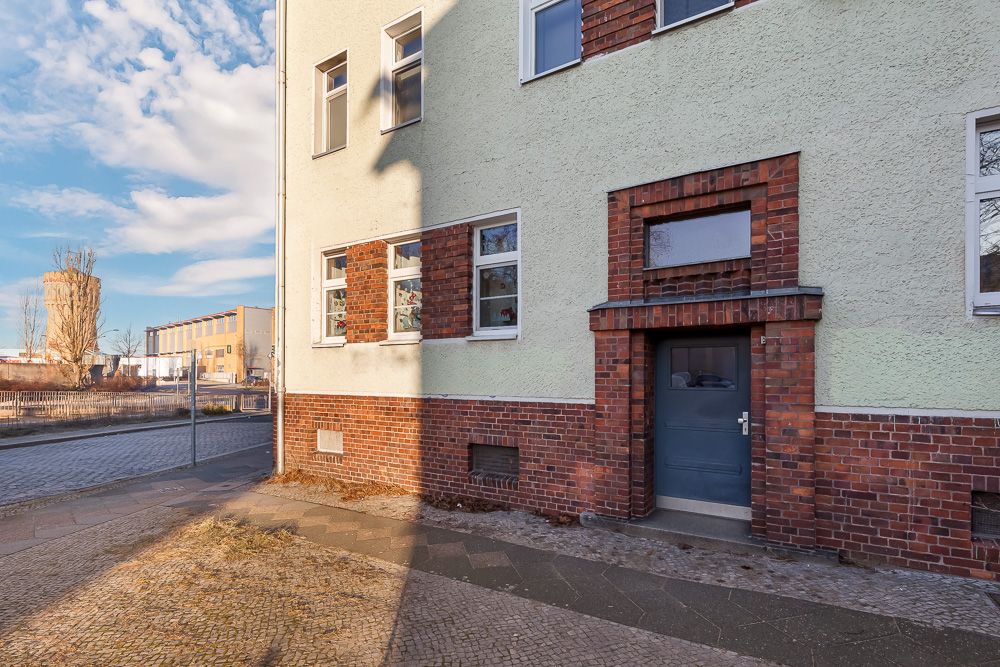 Eingangstür eines Mehrfamilienhauses mit Backsteinsockel und mehreren Fenstern an der Straße.