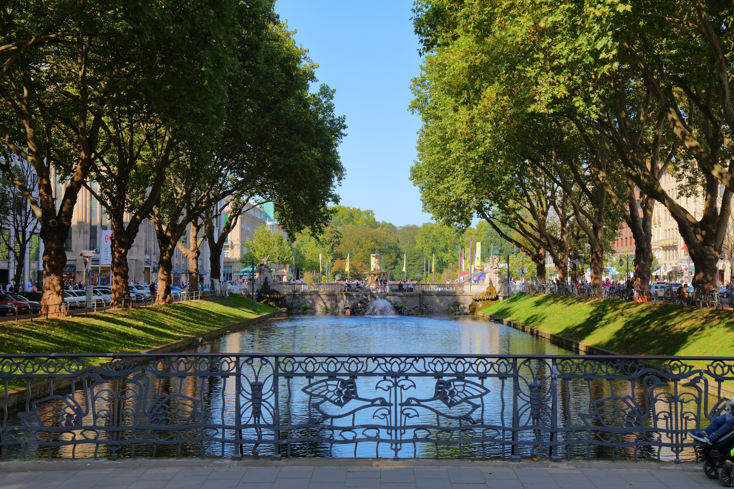 Kanal mit schmiedeeisernem Geländer, Bäumen und Brücke im Hintergrund bei sonnigem Wetter.