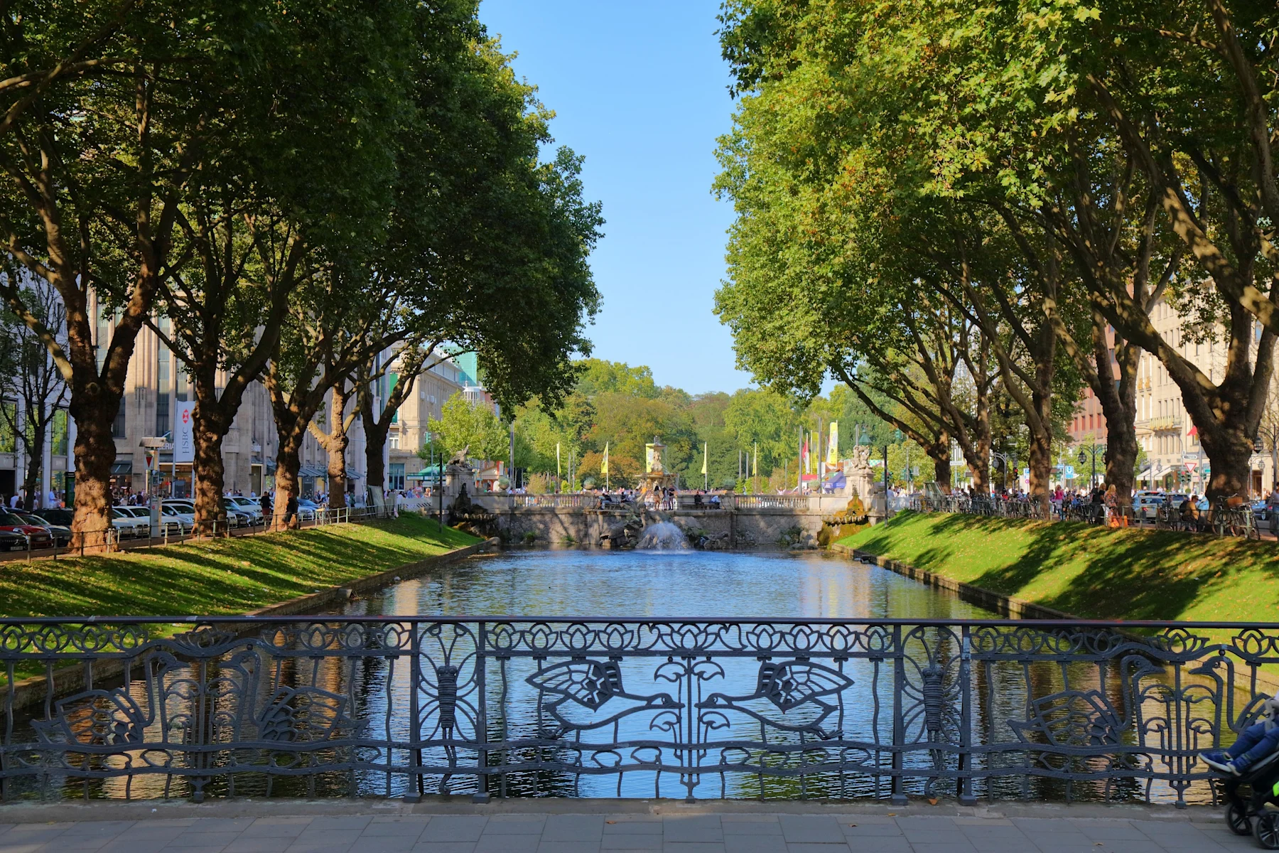 Kanal mit schmiedeeisernem Geländer, Bäumen und Brücke im Hintergrund bei sonnigem Wetter.