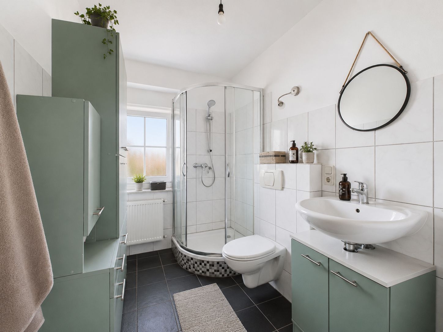 Helles Badezimmer mit Dusche, Waschbecken, WC, grünem Schrank und rundem Spiegel.