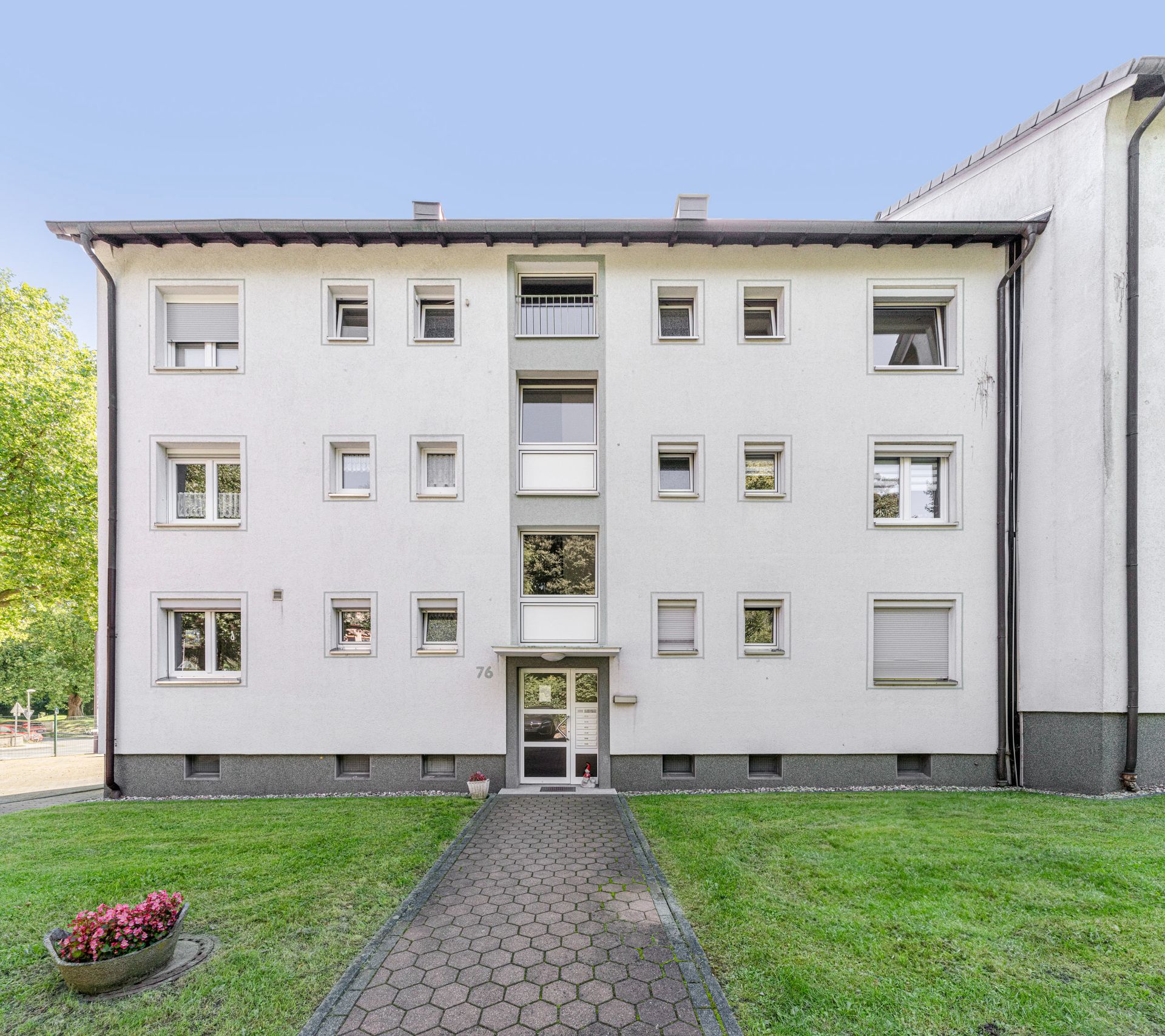 Drei-stöckiges Wohnhaus mit weißer Fassade und mehreren Fenstern, davor Rasen und Weg.