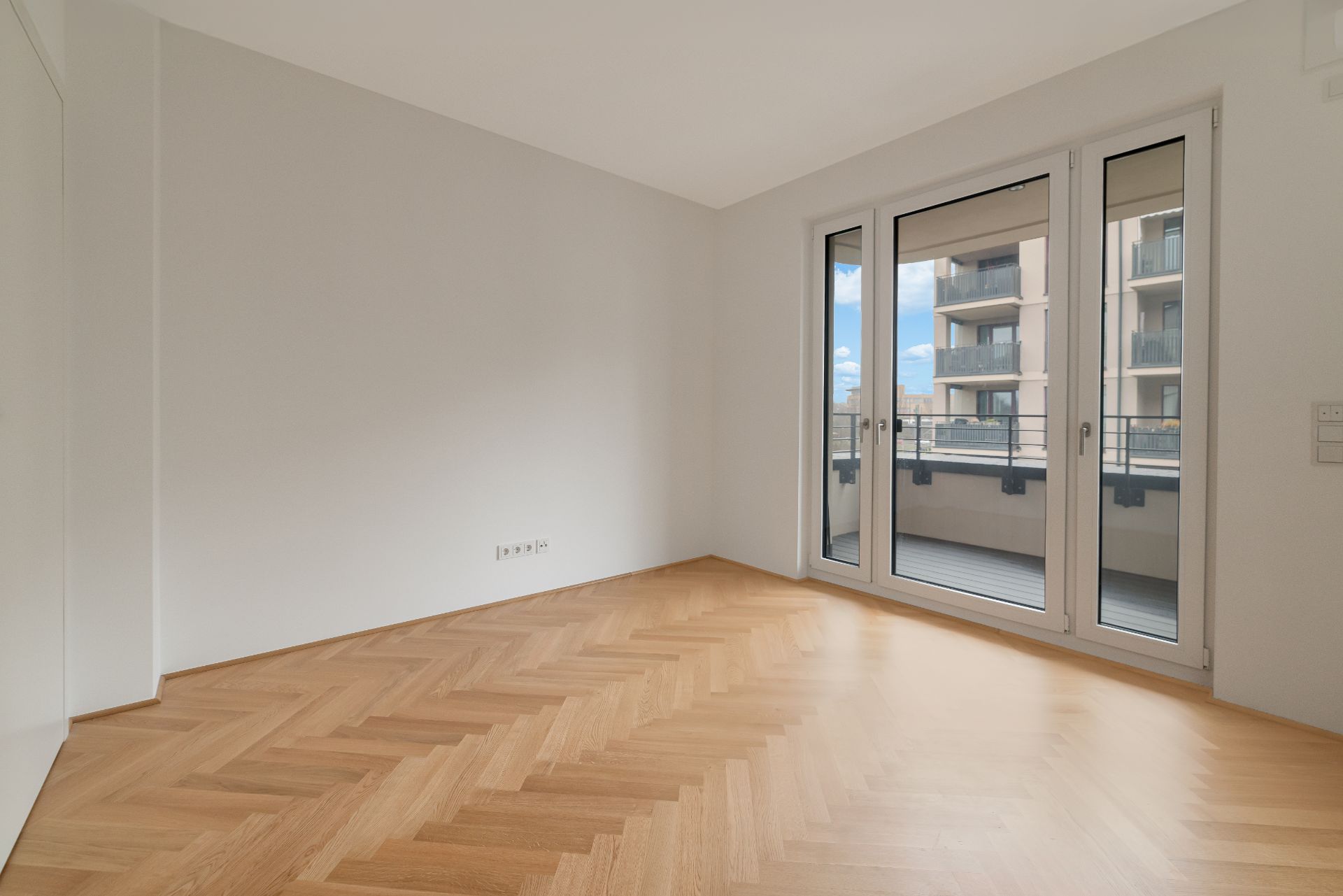 Helles, leeres Zimmer mit Parkettboden und Balkonzugang durch große Fenster.