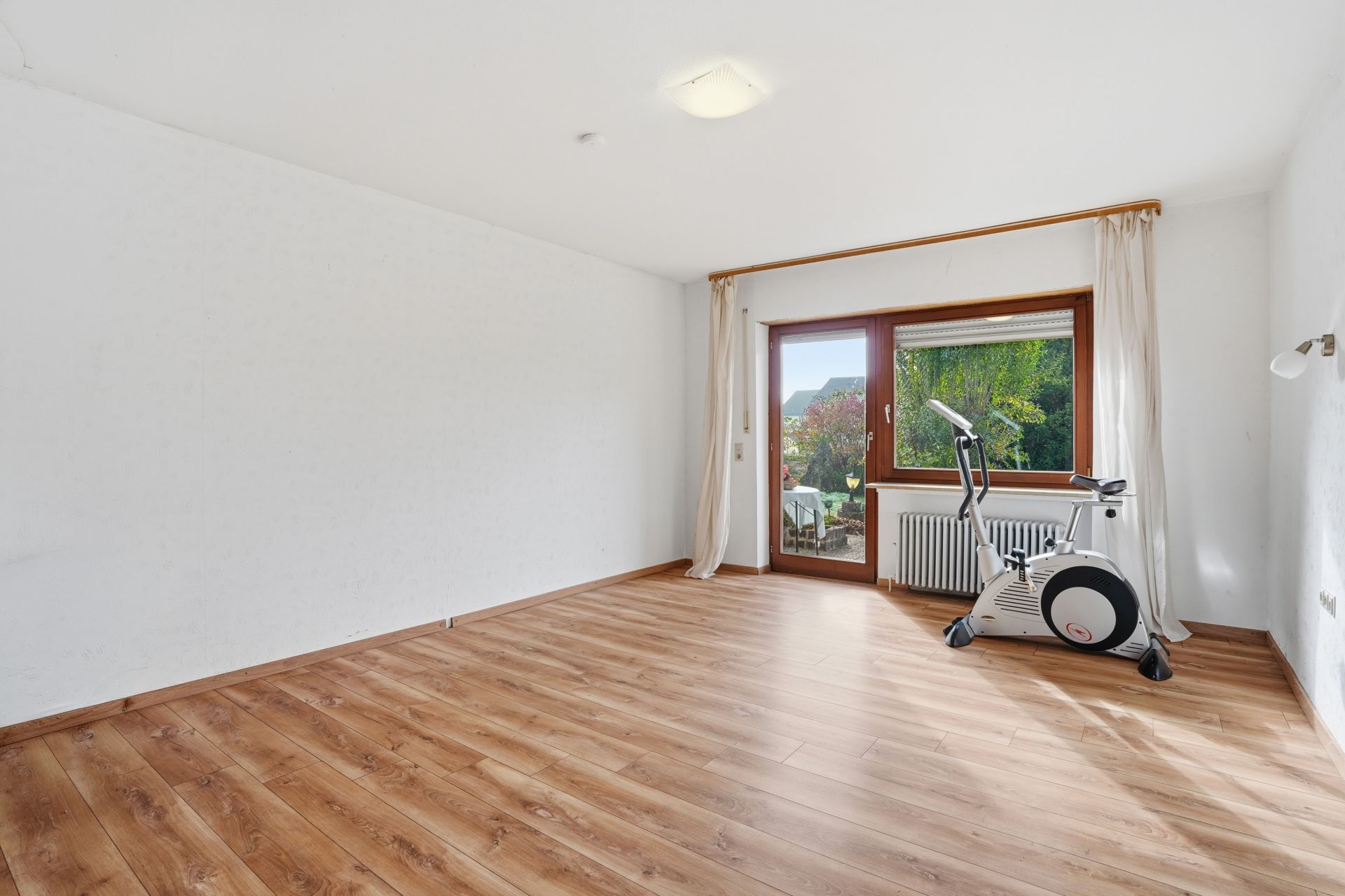 Helles Zimmer mit Holzfußboden, großem Fenster, Vorhängen und einem Heimtrainer am Fenster.