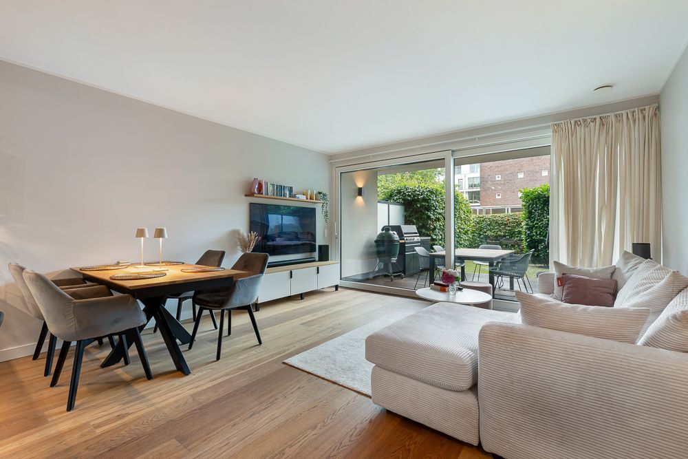 Modernes Wohnzimmer mit Sofa, Esstisch, Fernseher und Zugang zur Terrasse mit Grill.