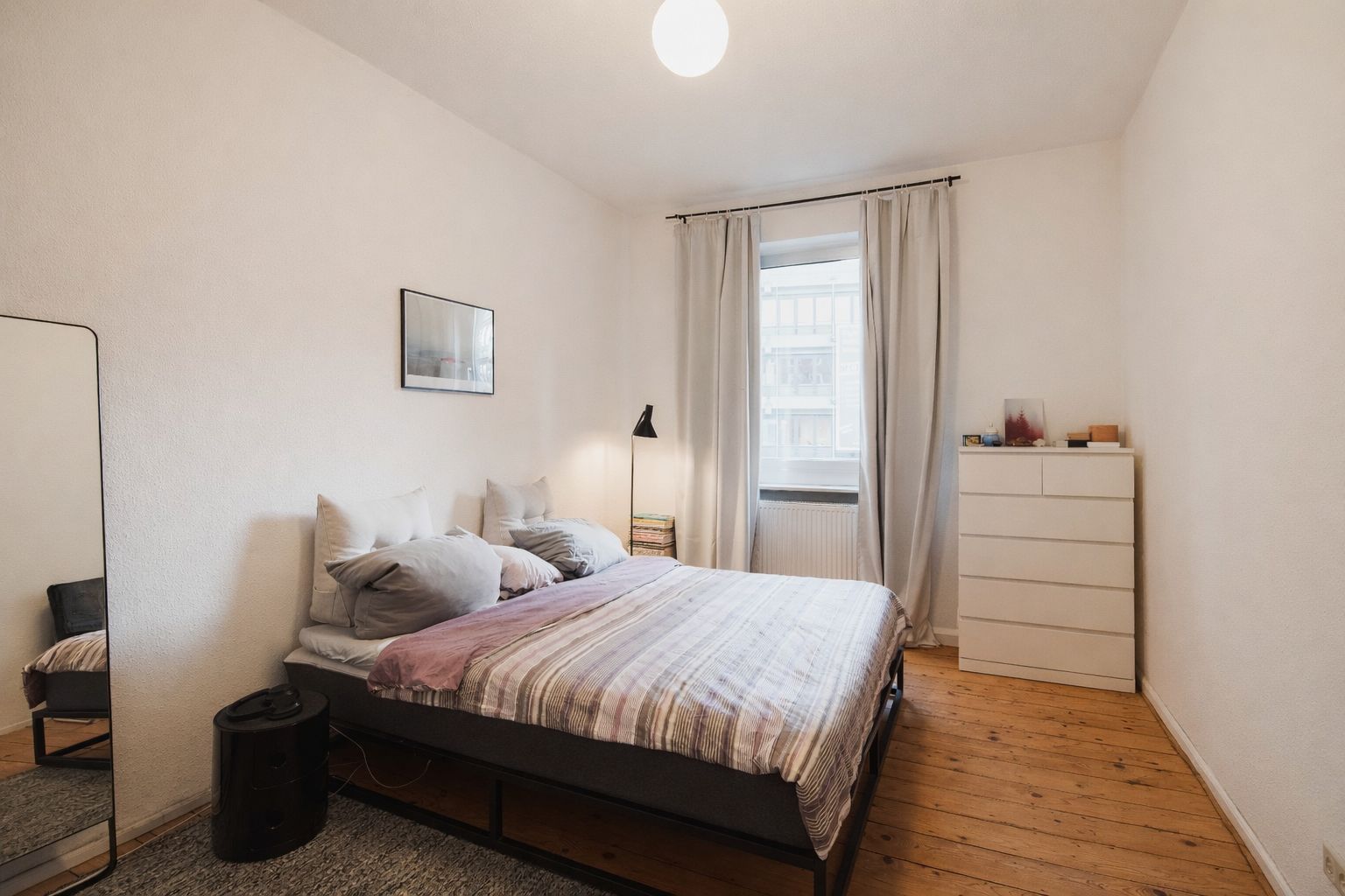 Schlafzimmer mit Doppelbett, Kommode, Spiegel und Fenster mit hellen Vorhängen.