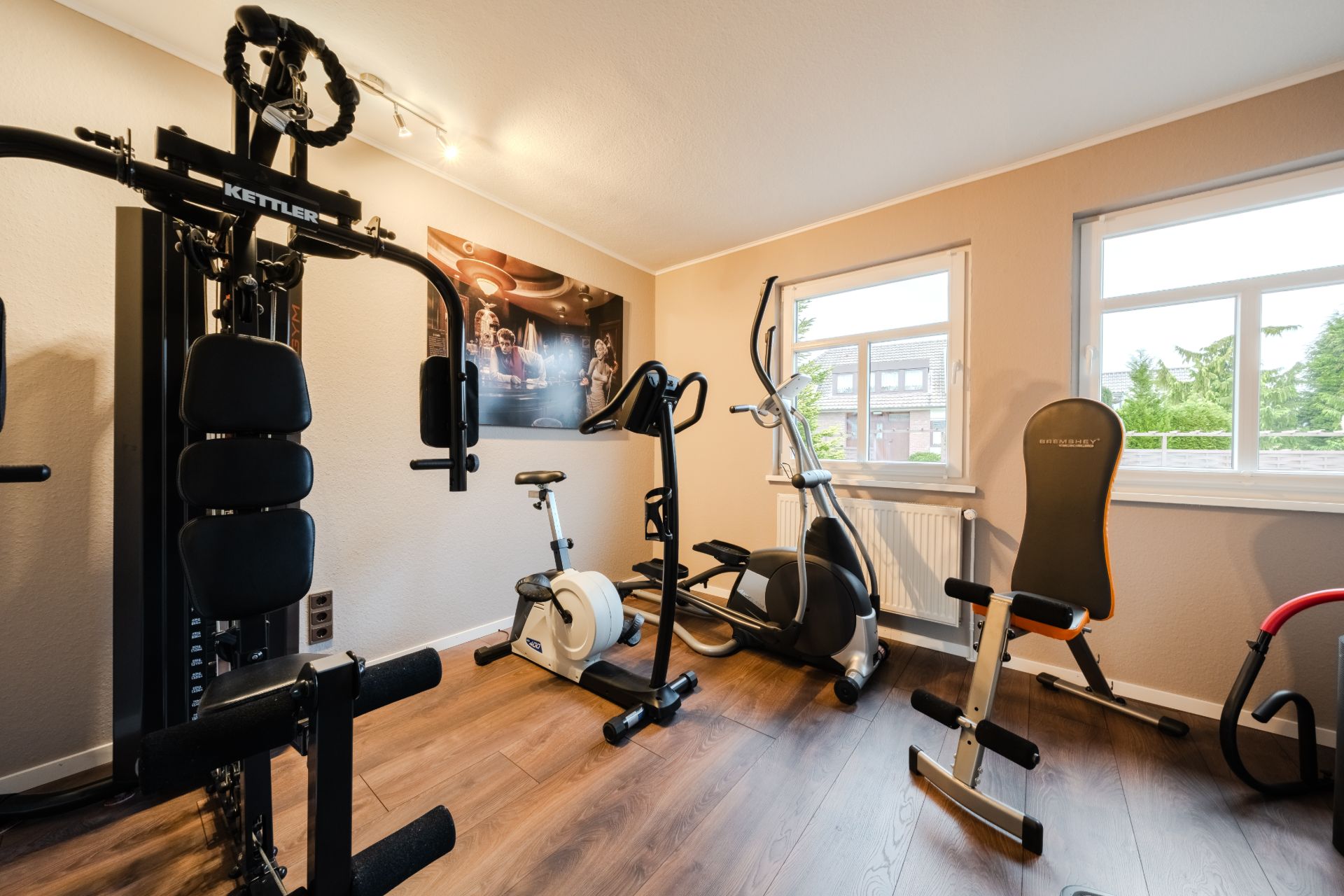 Heimfitnessraum mit verschiedenen Trainingsgeräten und Holzfußboden, zwei Fenster an der Wand.