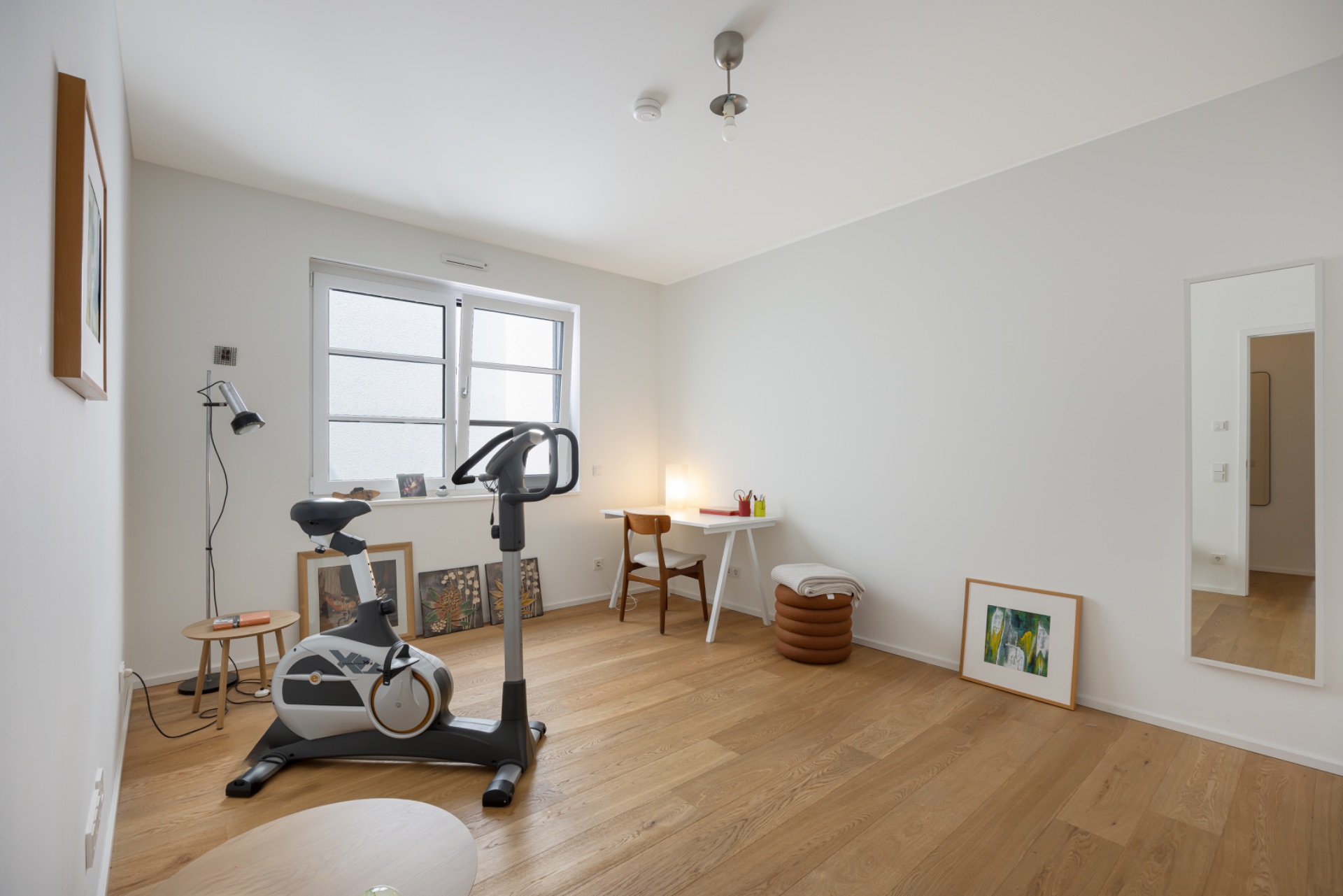 Helles Zimmer mit Heimtrainer, Schreibtisch, Holzfußboden und Bildern an den Wänden.