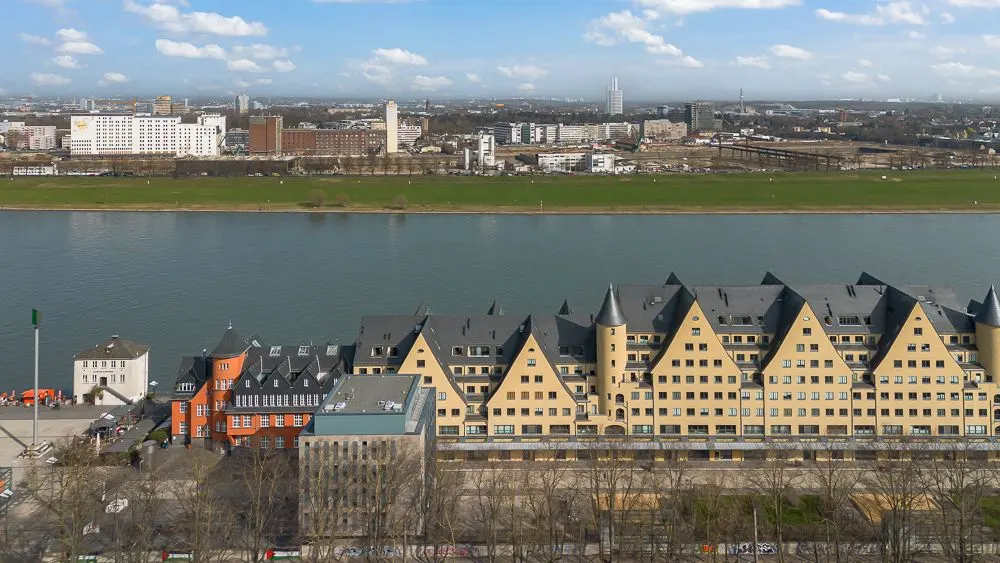 Blick auf Wohngebäude mit markanten Dächern am Flussufer, dahinter breite Wiese und Stadtlandschaft.