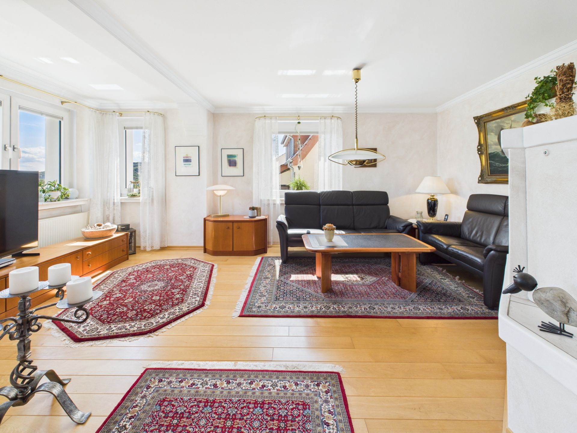 Helles Wohnzimmer mit zwei schwarzen Ledersofas, Holztisch und mehreren Teppichen auf Parkettboden.