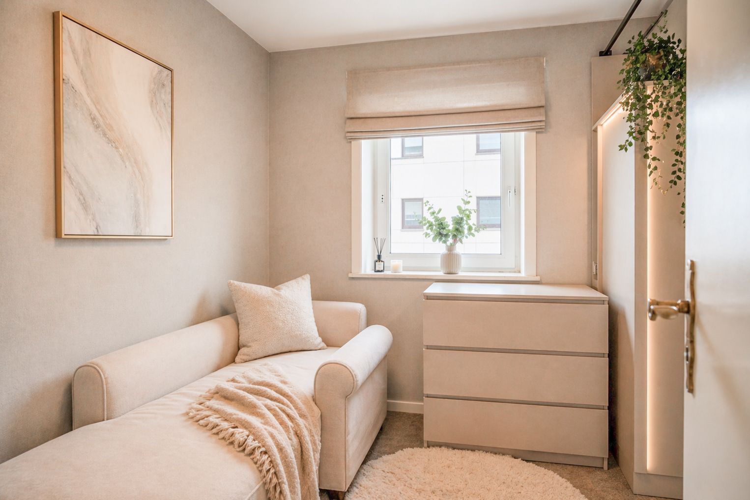 Helles Zimmer mit Sofa, Kommode, Fenster und Pflanze auf der Fensterbank.