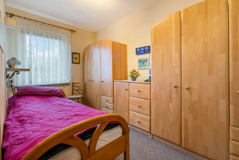 Kleines Schlafzimmer mit Einzelbett, Holzschränken und Fenster mit Gardine.