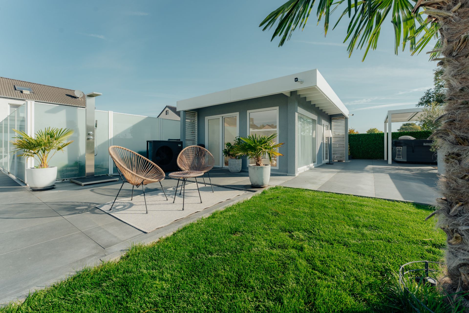 Moderne Terrasse mit zwei Stühlen, Rasen, Palmen und kleinem Gebäude im Hintergrund.