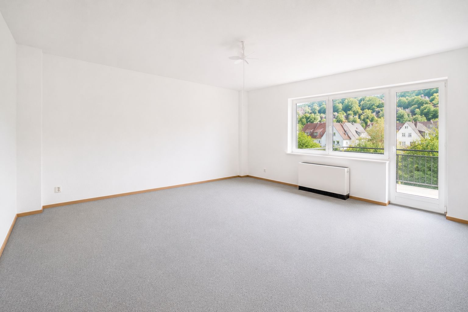 Helles, leeres Zimmer mit Teppichboden und großem Fenster mit Blick auf Häuser und Bäume.